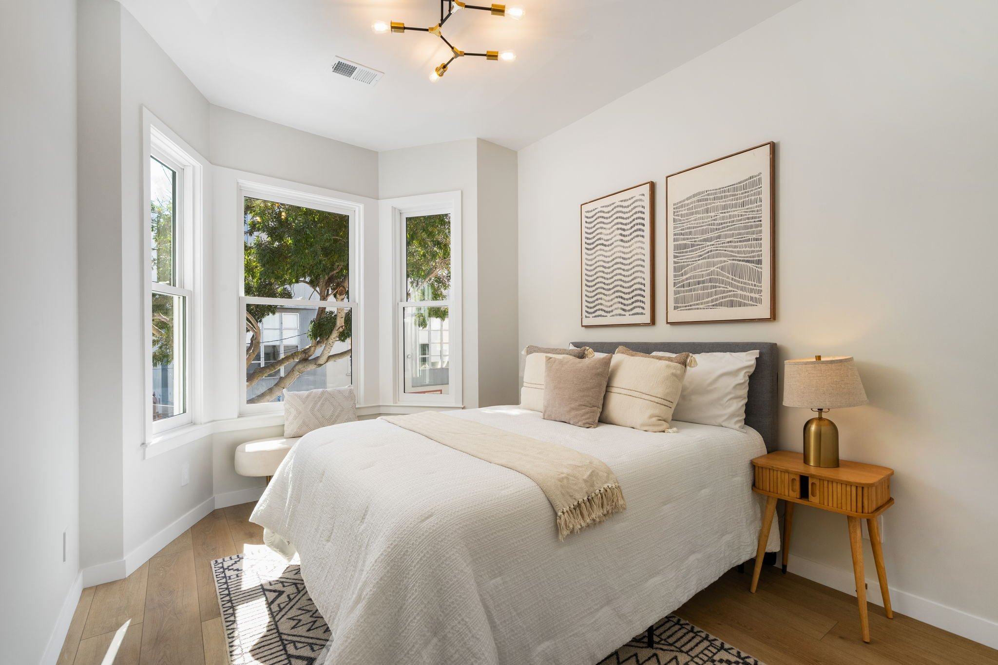 A bright, modern bedroom with a large bed covered in neutral-colored linens and several pillows. A wooden nightstand with a brass lamp is beside the bed. The room has white walls, a white ceiling, and a set of three large windows with views of green 