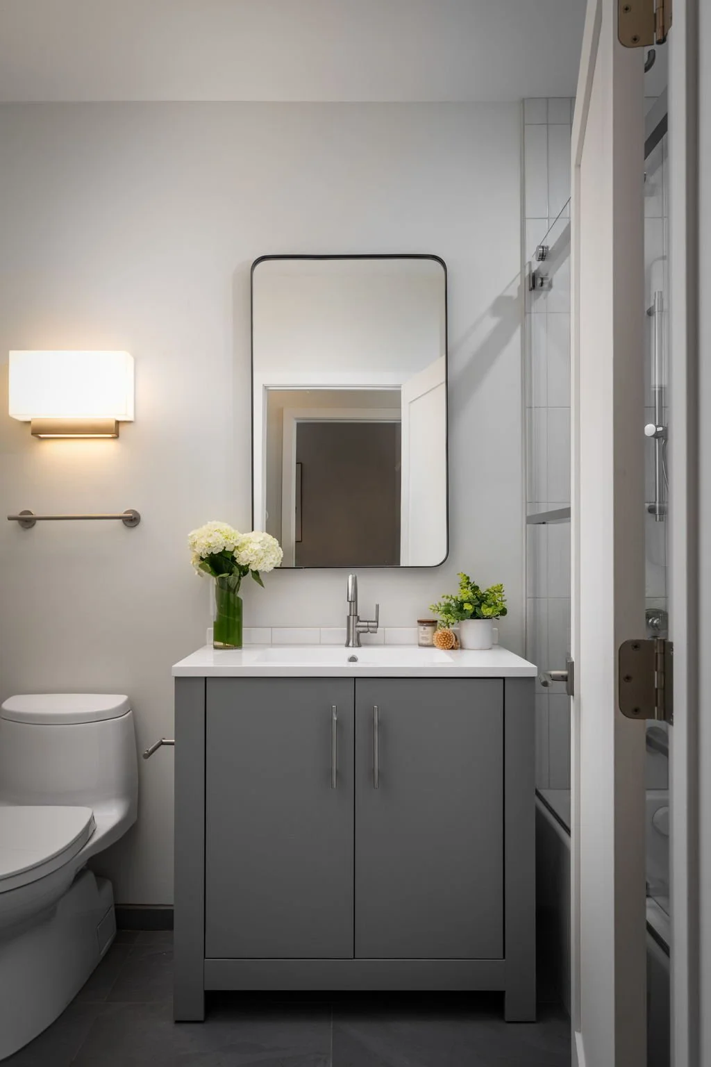 Modern bathroom with a gray vanity, white countertop, large mirror, flowers in a green vase, and a shower stall to the right.