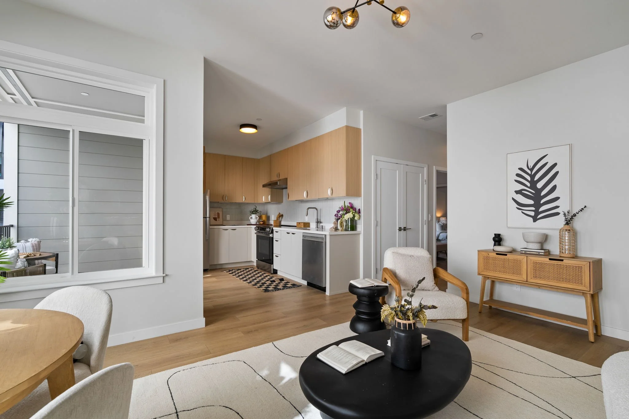 Open-concept living room and kitchen area featuring modern decor, light wood flooring, and white walls. The living space includes a black round coffee table with an open book on it, cream-colored chairs, and a wooden sideboard with decorative items. 