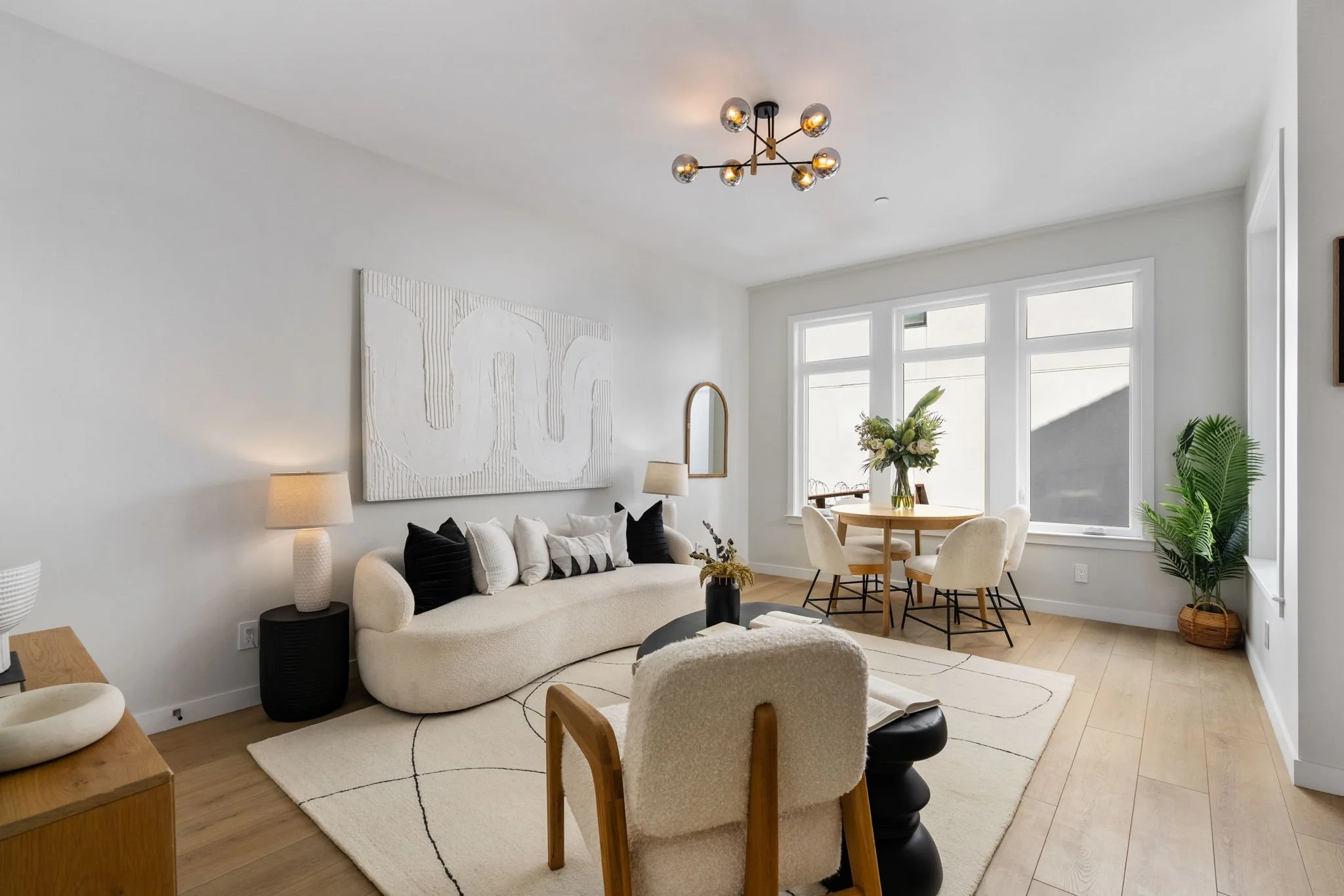 Bright living room with white walls, a white sofa, black and white pillows, a wooden side table, and a black round coffee table. There are large windows with white frames, a round wooden dining table with beige chairs, a tall green plant, and decorat