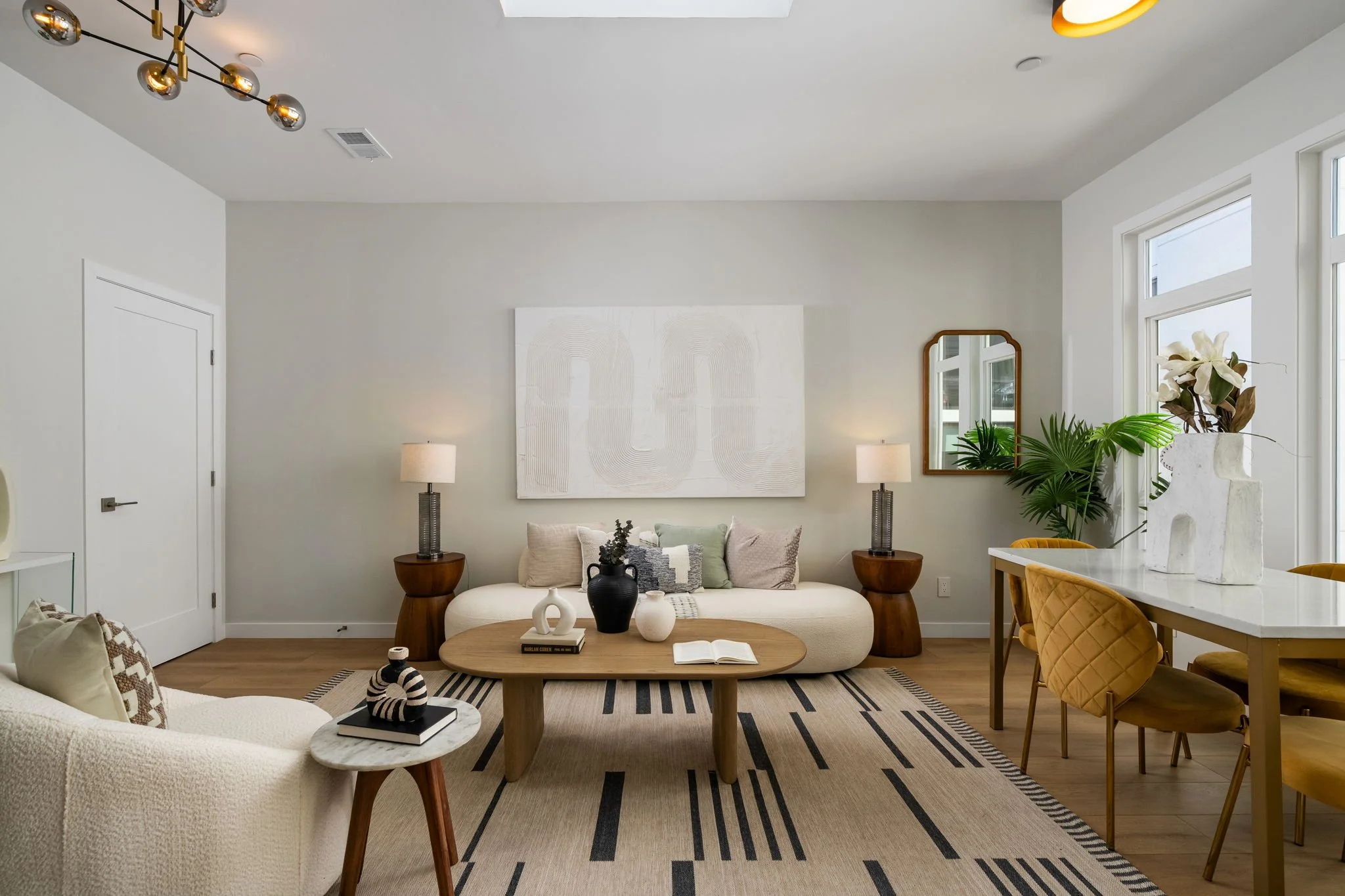 A contemporary living room with a white sofa, wooden coffee table, and various decorative items including vases, books, and pillows. There are two side lamps on either side of the sofa, plants near the windows, and a large abstract white artwork on t