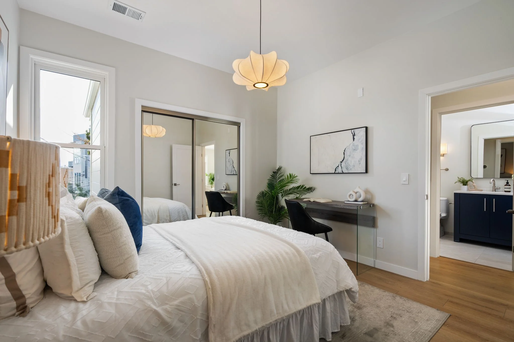 A bedroom with white walls, a large bed with white bedding and multiple pillows, a floating desk with a plant and decorative items, a black chair, artwork on the wall, a window with sunlight, and a bathroom visible through an open door.
