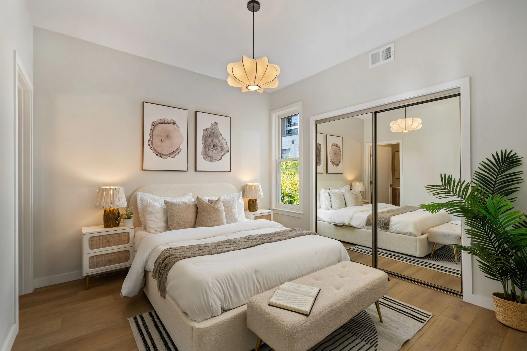 A modern bedroom with a large bed featuring white and beige bedding, surrounded by two bedside tables with matching lamps, artwork on the walls, a mirrored closet, and a tall potted plant, with natural light coming through a window.