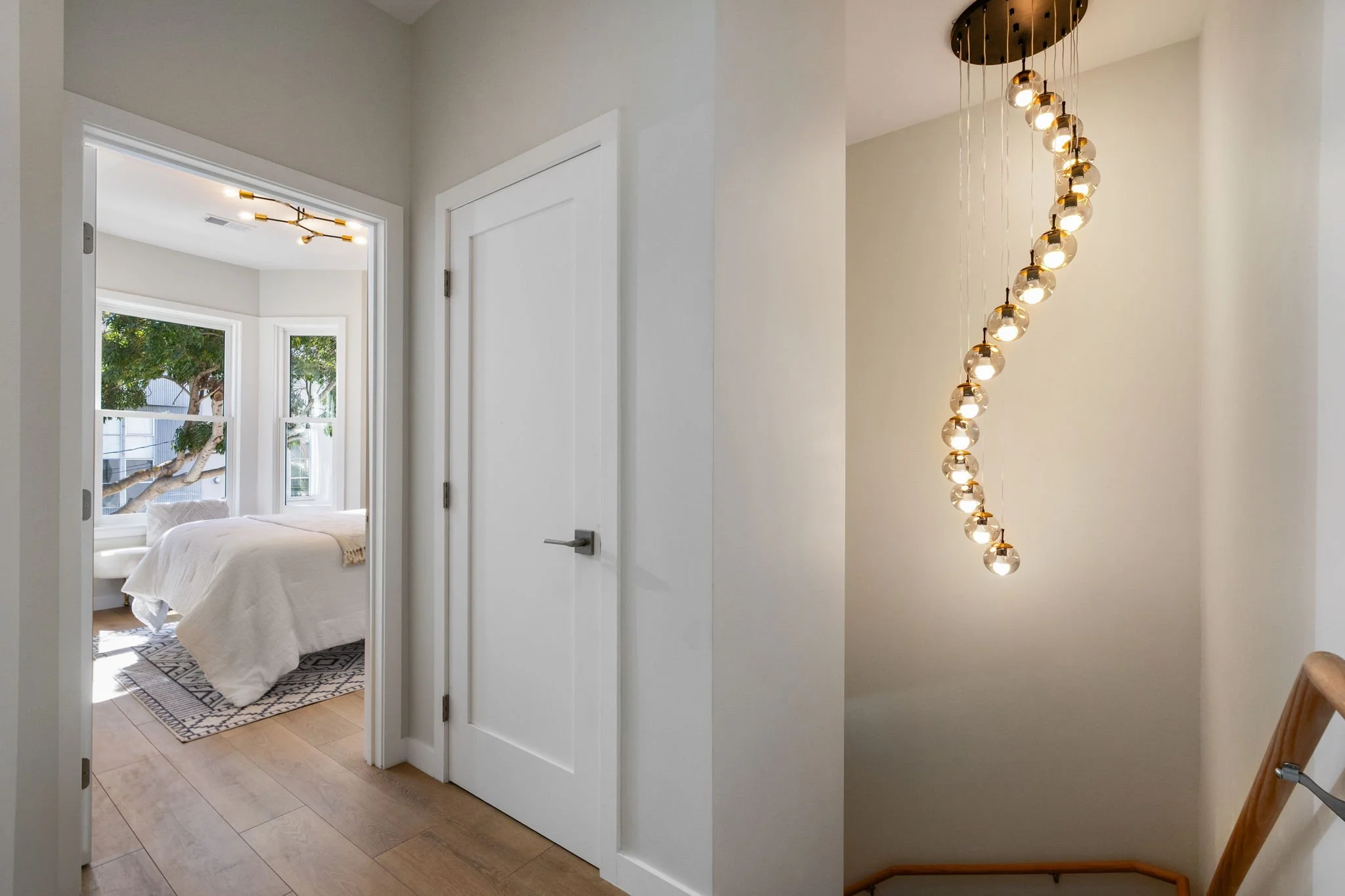 Interior view showing a hallway leading to a bedroom with a large window and white bedding, a modern chandelier with multiple spherical lights hanging in the hallway, and a wooden staircase with a handrail on the right side.