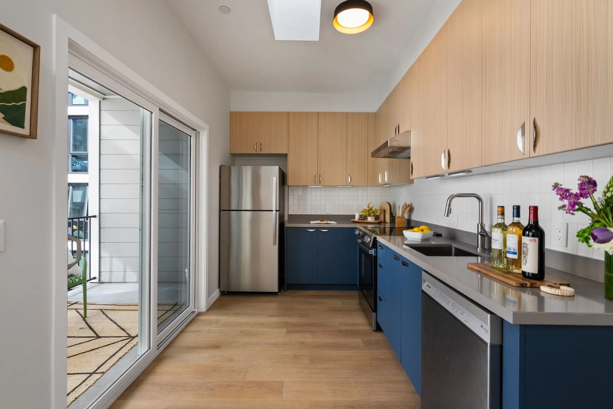 A modern kitchen with light wood cabinets, dark blue lower cabinets, stainless steel refrigerator, white tile backsplash, and a white countertop with wine bottles, a green plant, and fresh flowers. There is a sliding glass door leading to a balcony o
