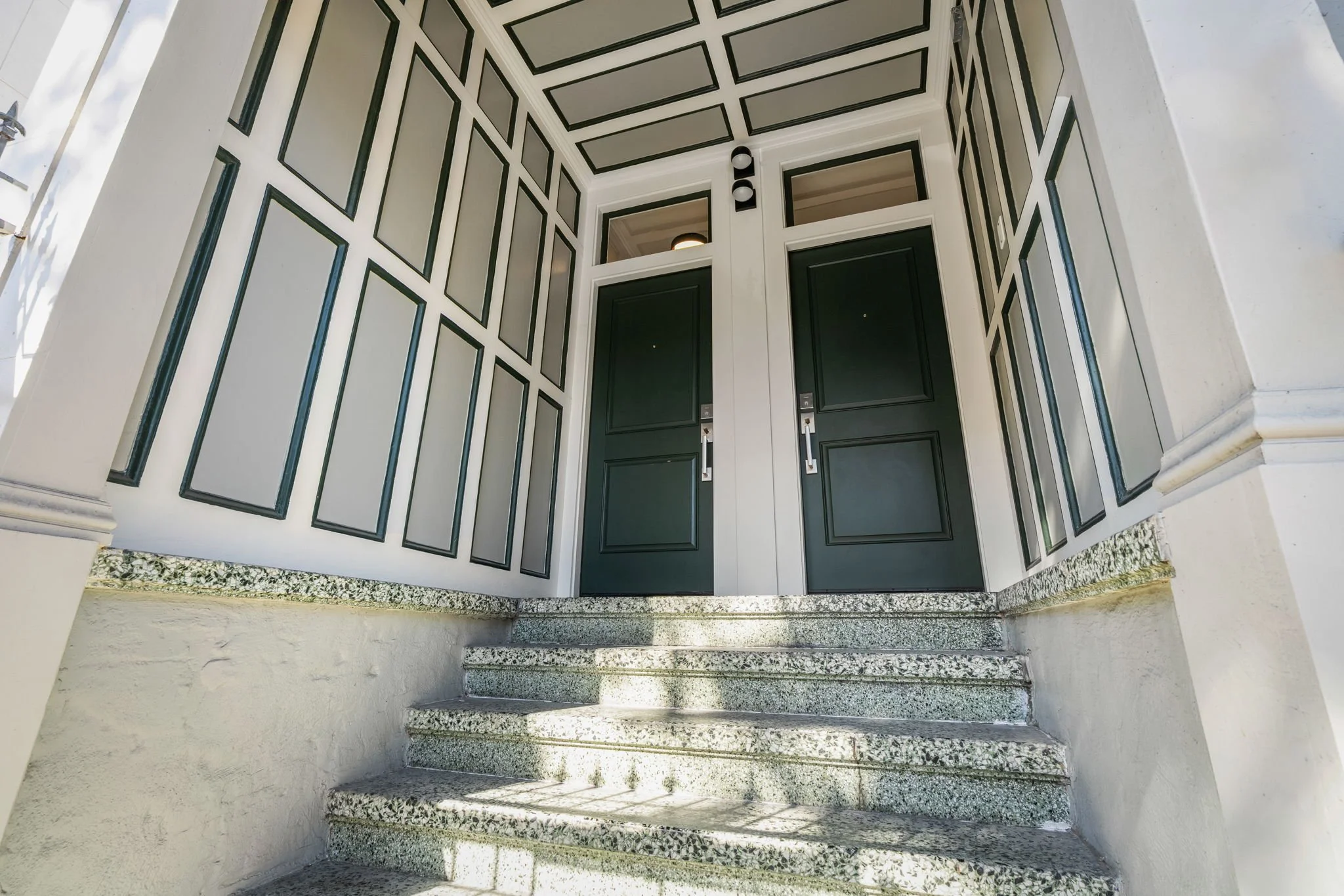 Exterior entrance of a building with a dark green door, framed by a white wall with black trim in a geometric pattern, and granite steps leading up to the door.