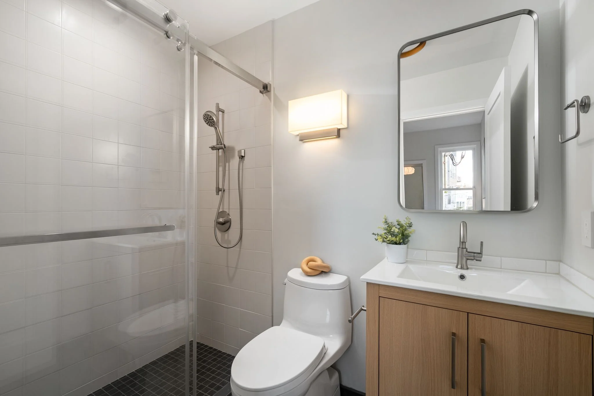 A modern bathroom with a white tiled walk-in shower, a toilet, a wooden vanity with a sink, a mirror, a wall light, and a window. There is a small potted plant and a towel on the toilet lid.