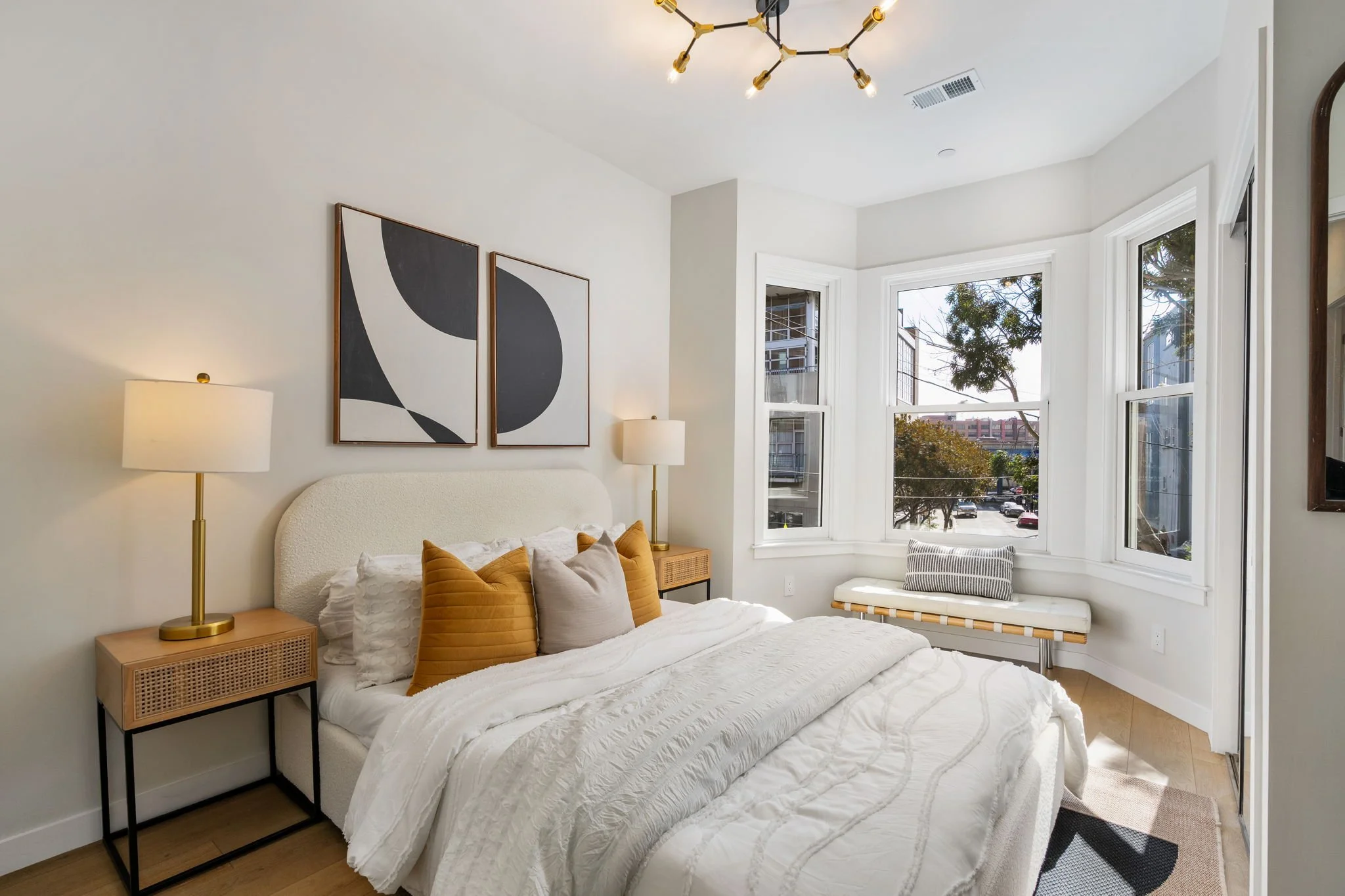 A cozy bedroom with a white bed, mustard yellow pillows, and a textured white comforter. Two gold desk lamps sit on matching bedside tables, and two framed abstract black and white art pieces adorn the wall above the bed. Large windows allow natural 