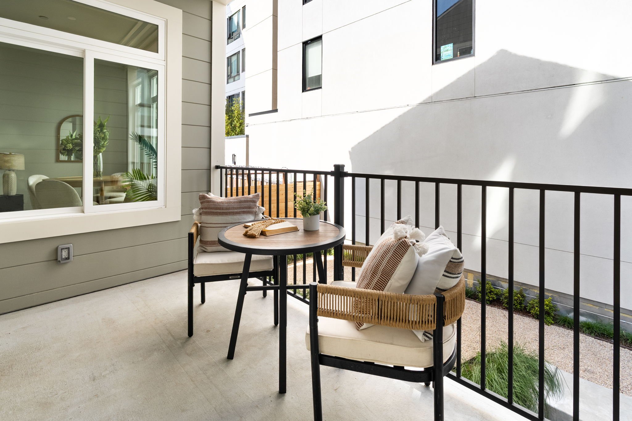 A small balcony with a black metal railing, outdoor seating including two chairs with cushions, a round black table with a potted plant and a book, adjacent to a large window showing an interior with a table, lamp, and houseplants, and neighboring mo