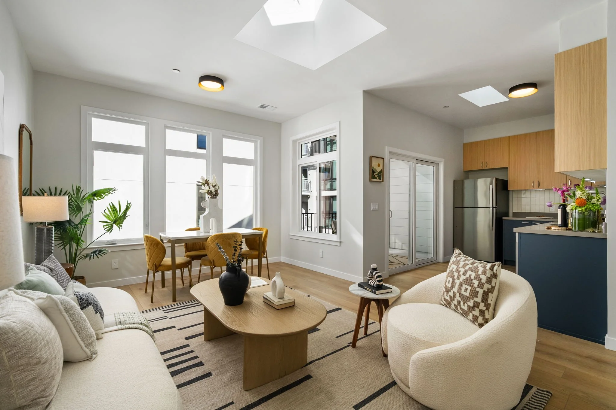 Living and dining area with white walls, large windows, wooden flooring, and modern light fixtures ceiling. There is a white sofa, armchair, a wooden coffee table with decorative vases, and a dining table with yellow chairs. The kitchen has wooden cabinets, a stainless steel refrigerator, and a colorful flower arrangement.