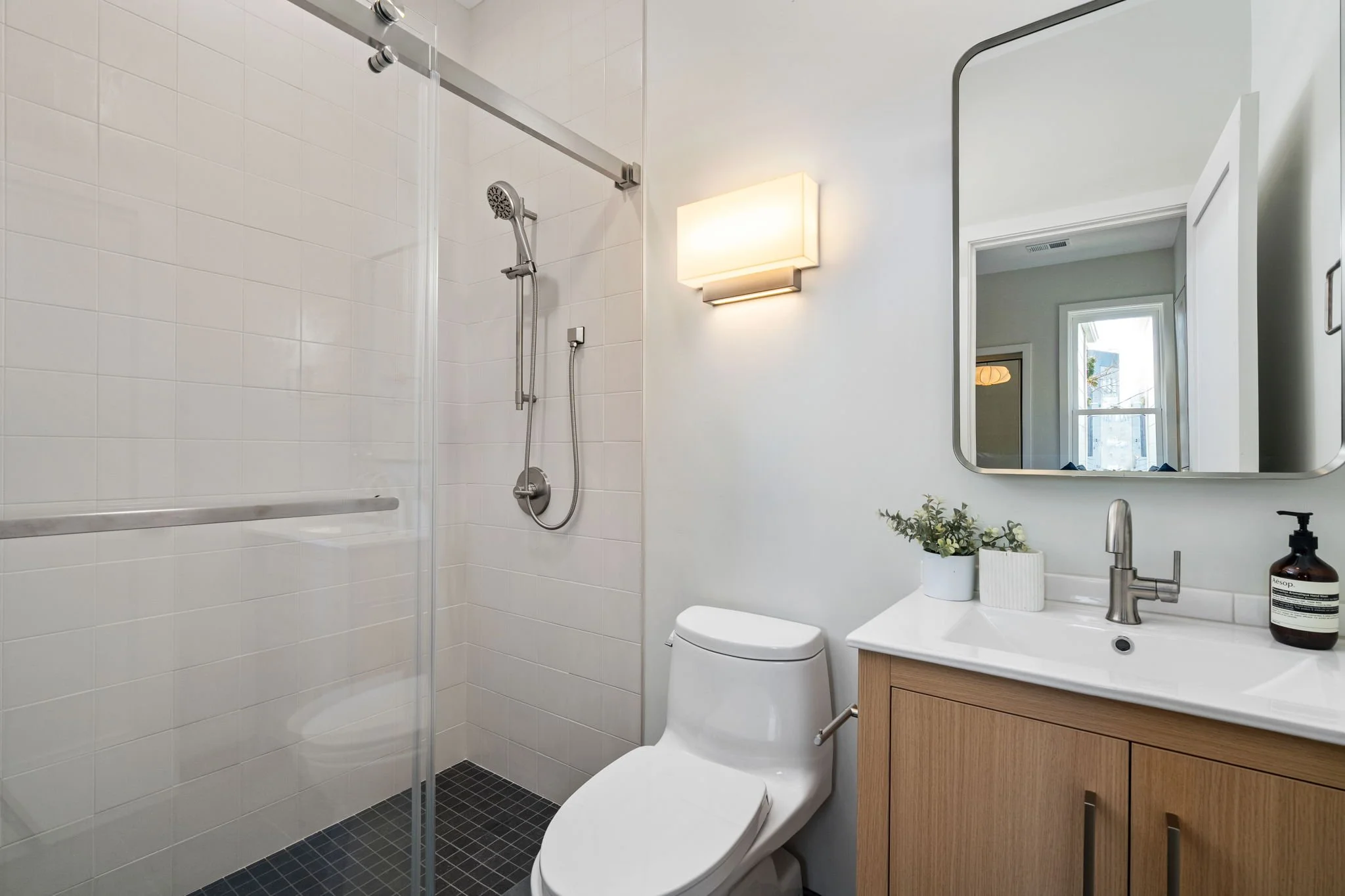 A bathroom with a glass shower enclosure, a wall-mounted rectangular light fixture, a mirror, a vanity with a sink, a potted plant, a bottle of soap, and a toilet.