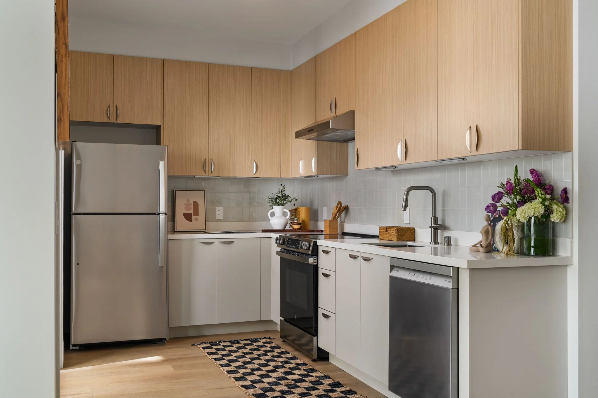 A modern kitchen with light wooden cabinets, stainless steel appliances including a refrigerator and stove, a white countertop, a sink with a faucet, and decorative items like a colorful bouquet of flowers, framed artwork, and a potted plant.