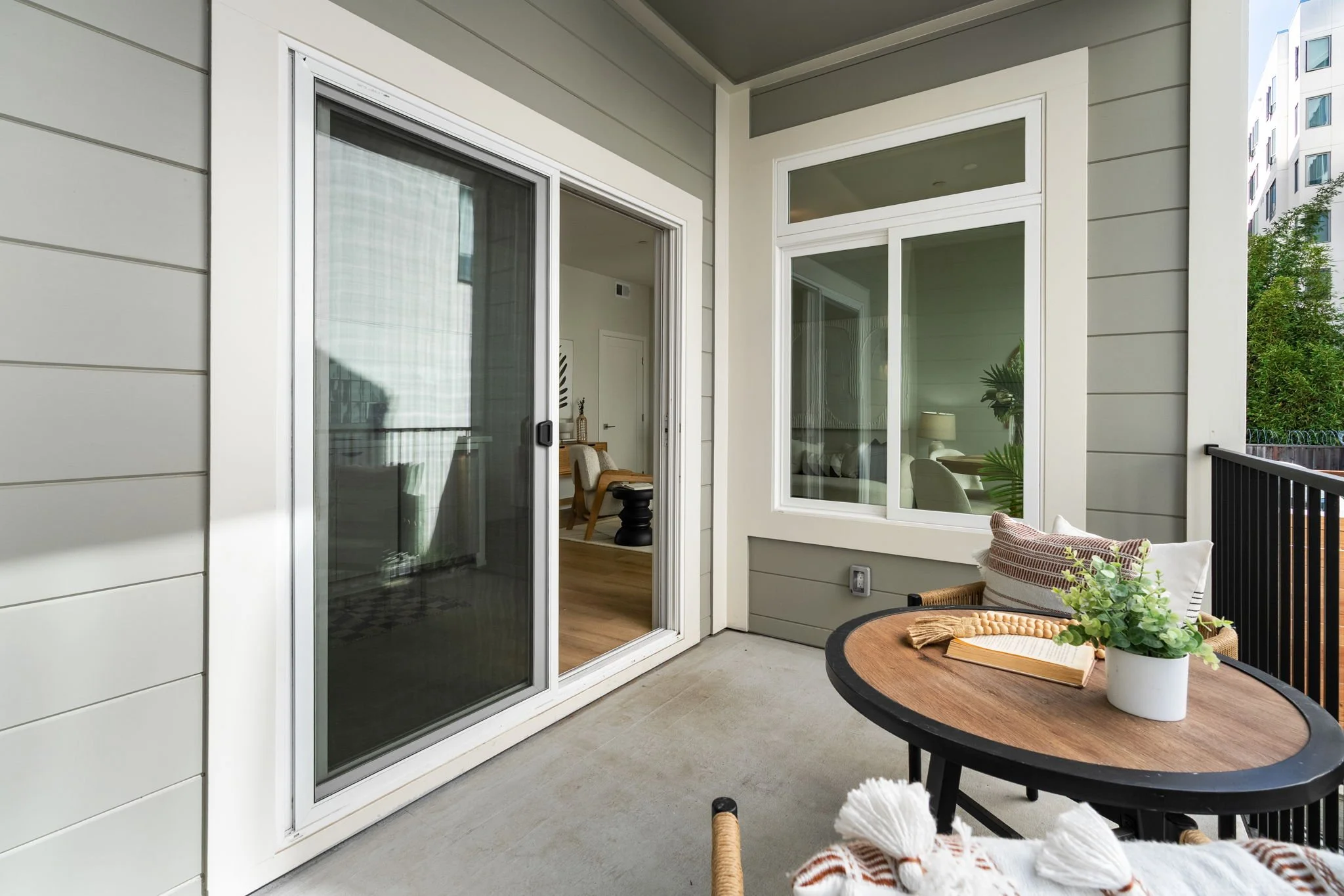 Balcony with sliding glass door, a small round table with a plant and a book, outdoor seating, and view of neighboring building.