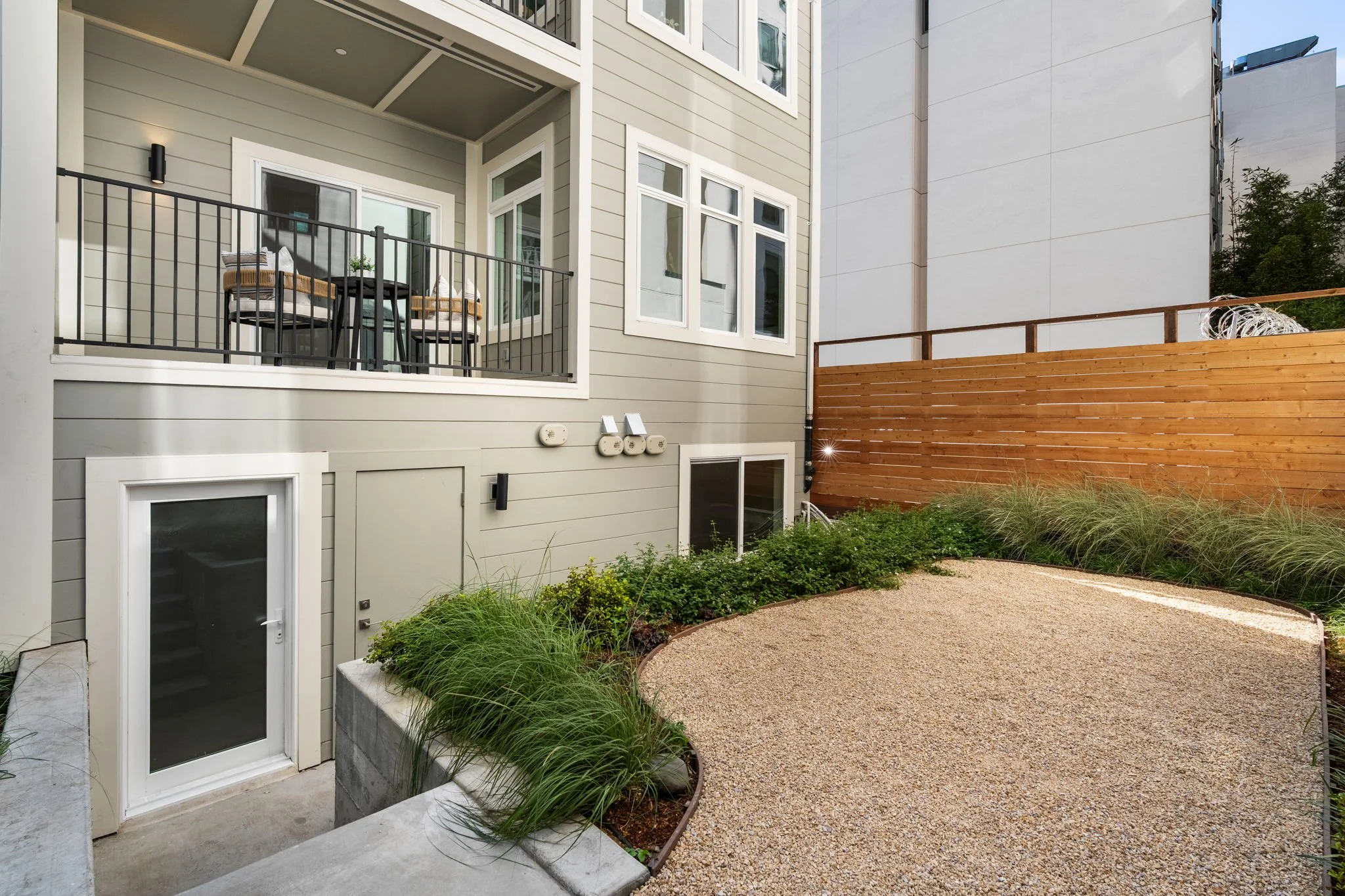 Backyard patio area with a small table and two chairs on a balcony, a garden with plants, grass, and a gravel pathway, a wooden fence, and a multi-story beige house with large windows.