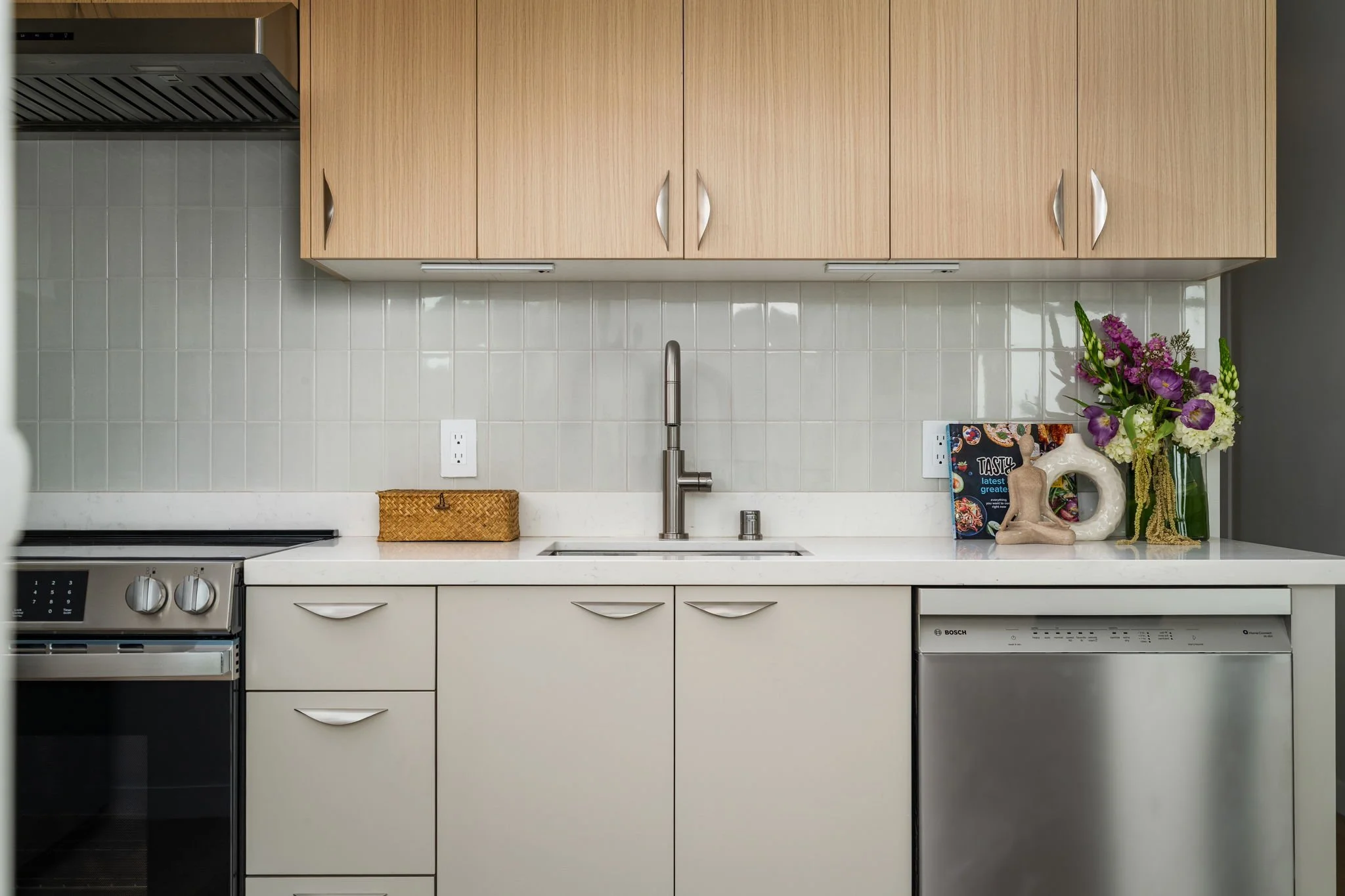 Modern kitchen with wooden upper cabinets, white lower cabinets, a white quartz countertop, a stainless steel sink with a gooseneck faucet, beige tiled backsplash, and decorative items including a bouquet of flowers and a small sculpture.
