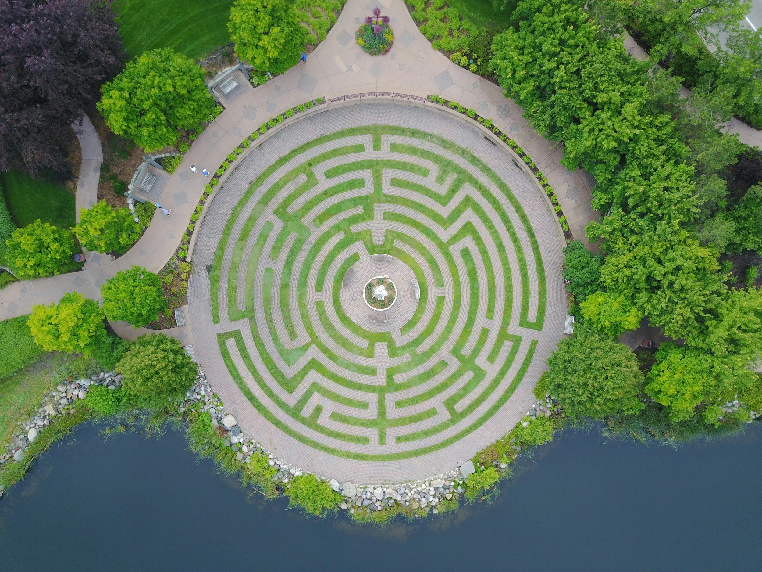Vue aérienne d'un jardin avec un labyrinthe en forme de cercle, entouré d'eau et de verdure, avec des arbres et des sentiers pavés.