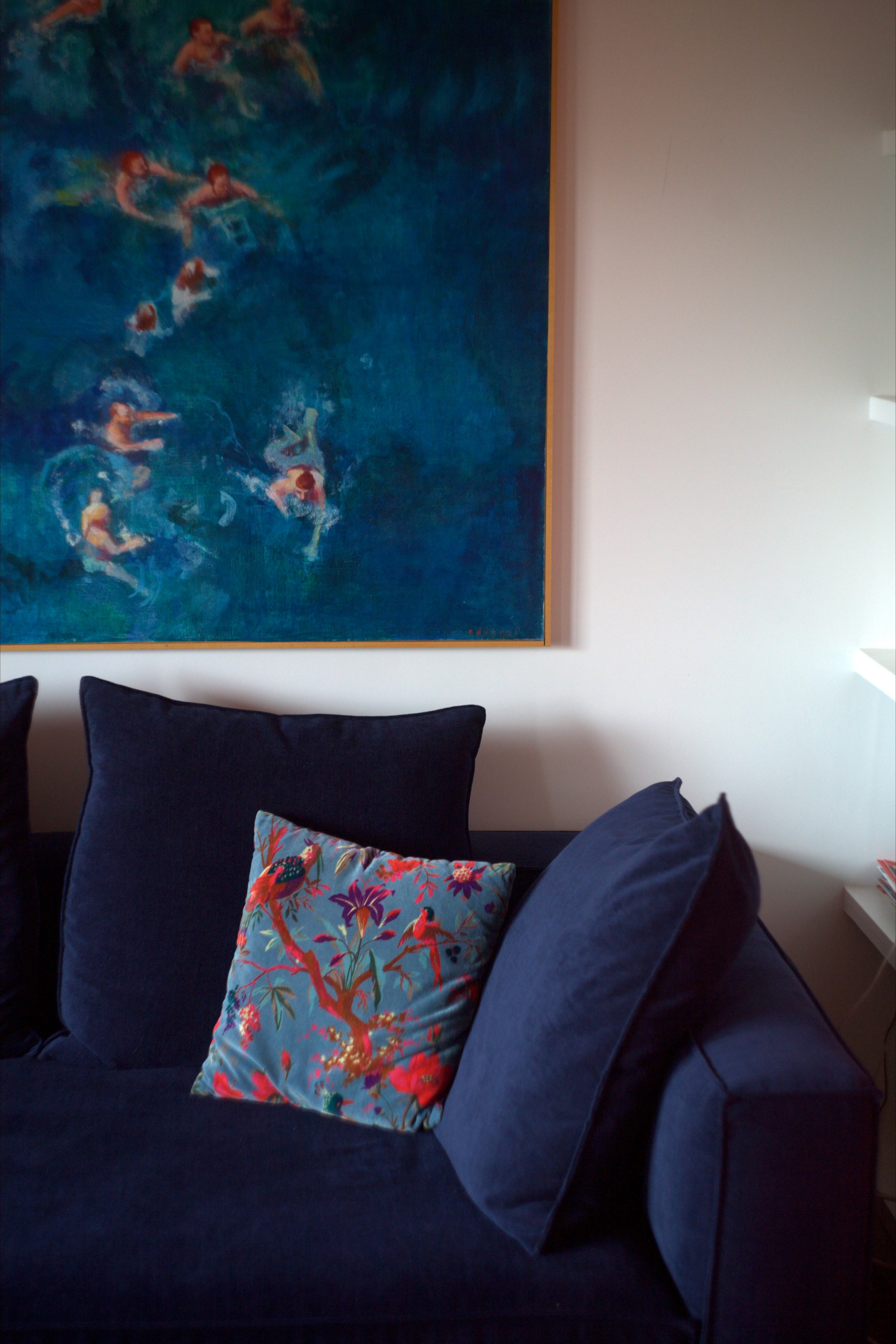 Salon avec un canapé noir, un coussin coloré à motif floral, un tableau représentant des nageurs en mer, et un mur blanc.