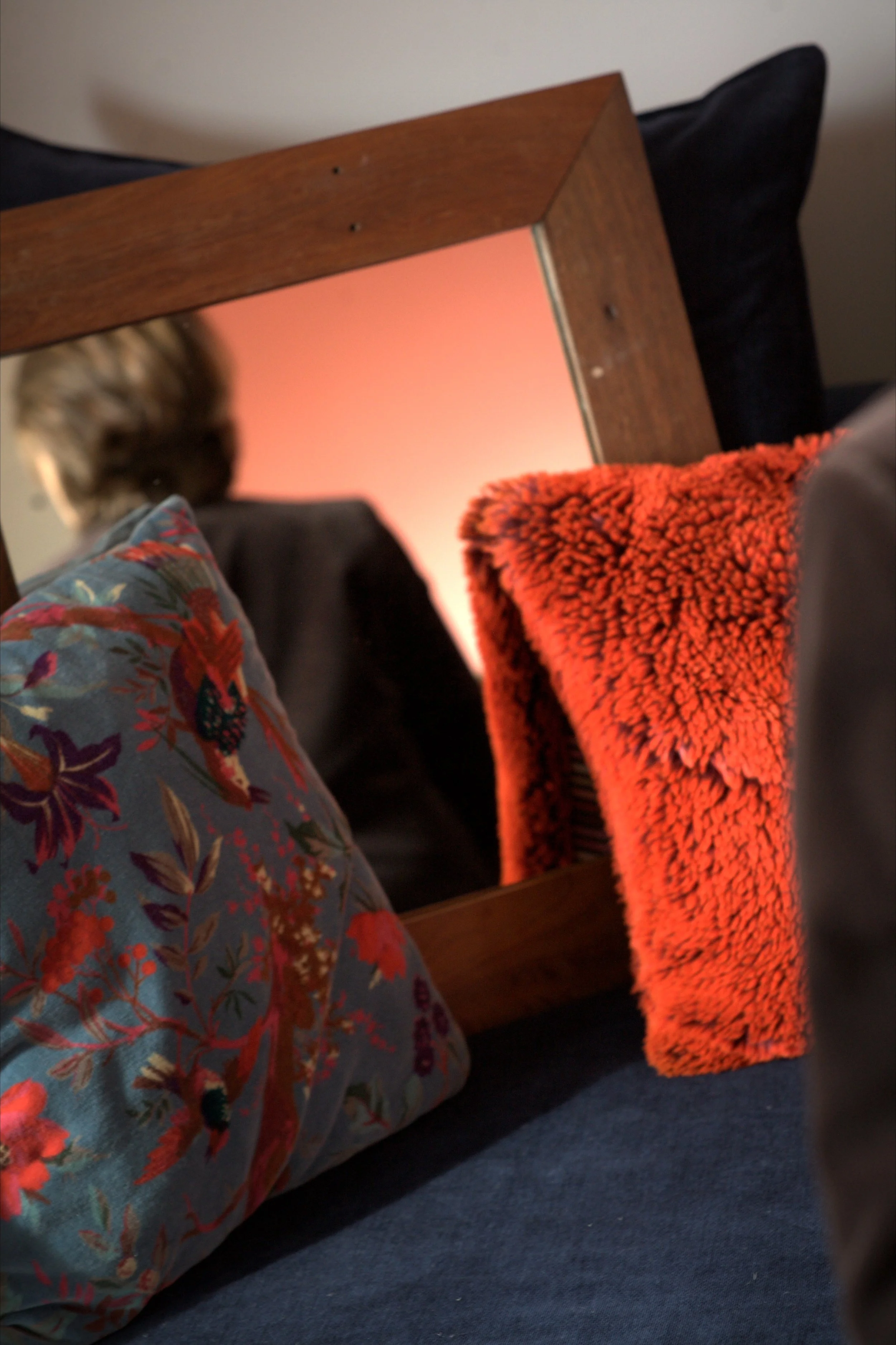 Un miroir placé sur une table en bois, reflétant un plafond orange et une personne portant un vêtement à motif floral. Sur la table, un coussin noir et une couverture orange peluche.