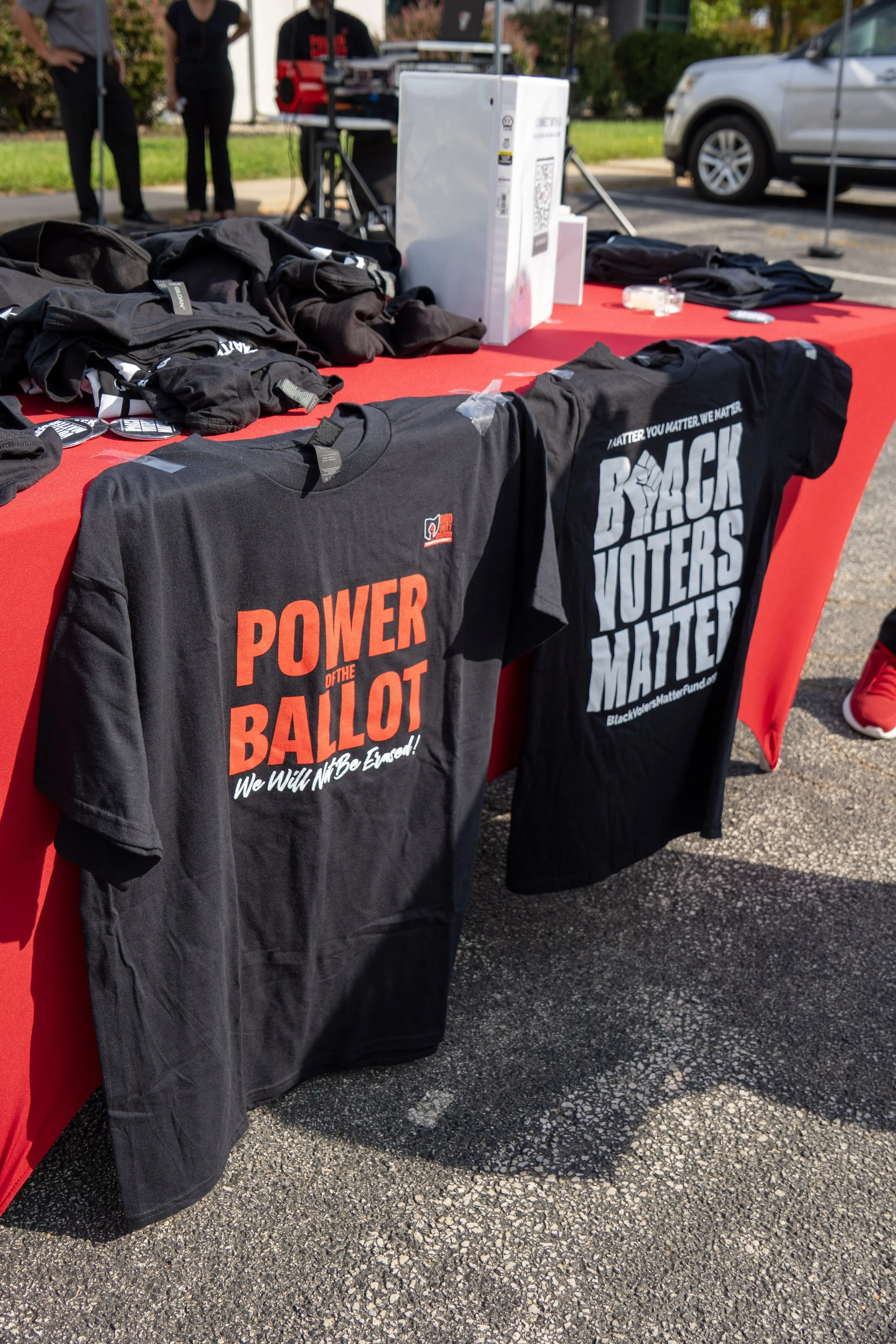 Table with black t-shirts promoting voting rights and political activism, set outdoors with people in the background.