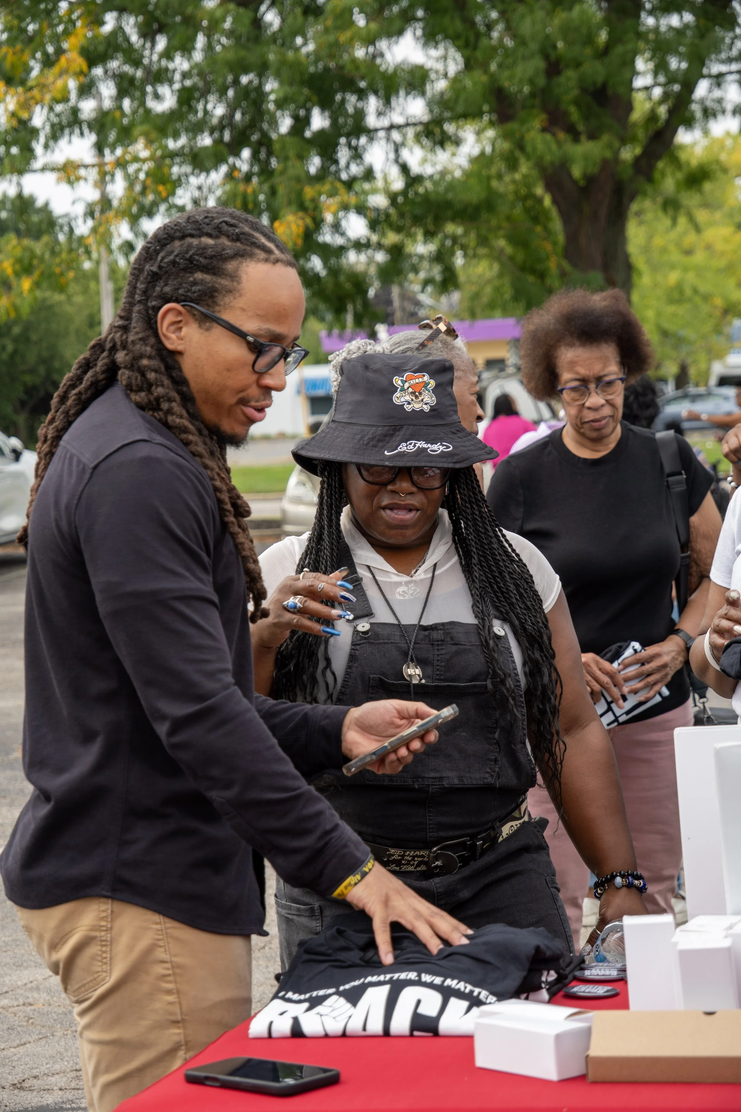 People gathered outdoors around a table with merchandise, including a black t-shirt with white and yellow text and a graphic, with a man and two women looking at items and talking.