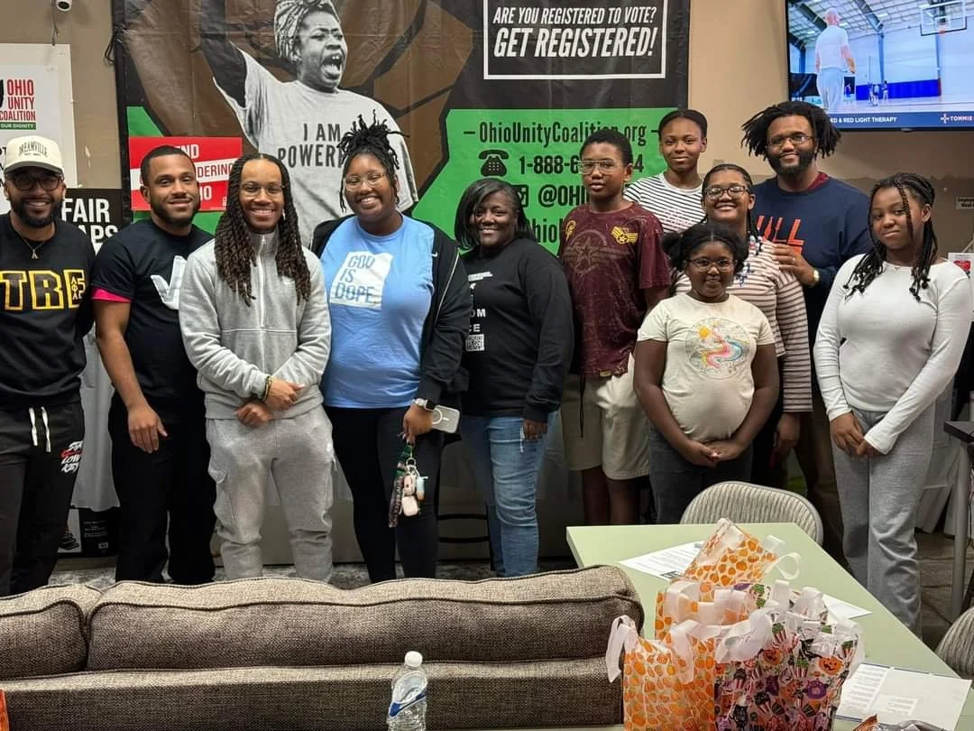Group of people standing together in front of a wall with signs and a large photo of a woman. The group includes men and women of different ages, some smiling and dressed casually. There is a table with wrapped gifts and a water bottle in the foreground.