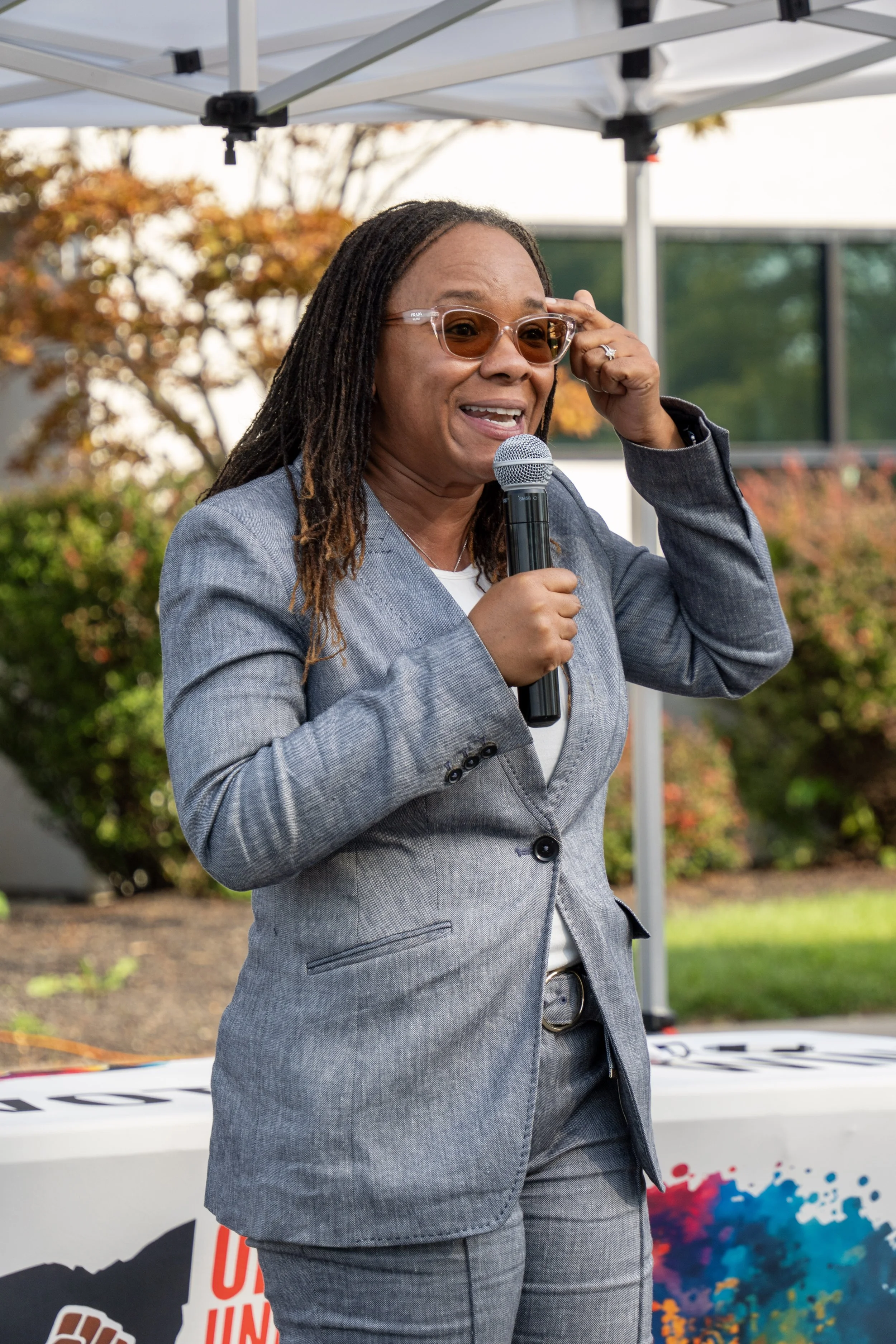 A woman with dreadlocks, wearing glasses and a gray suit, speaking into a microphone outdoors during daytime.
