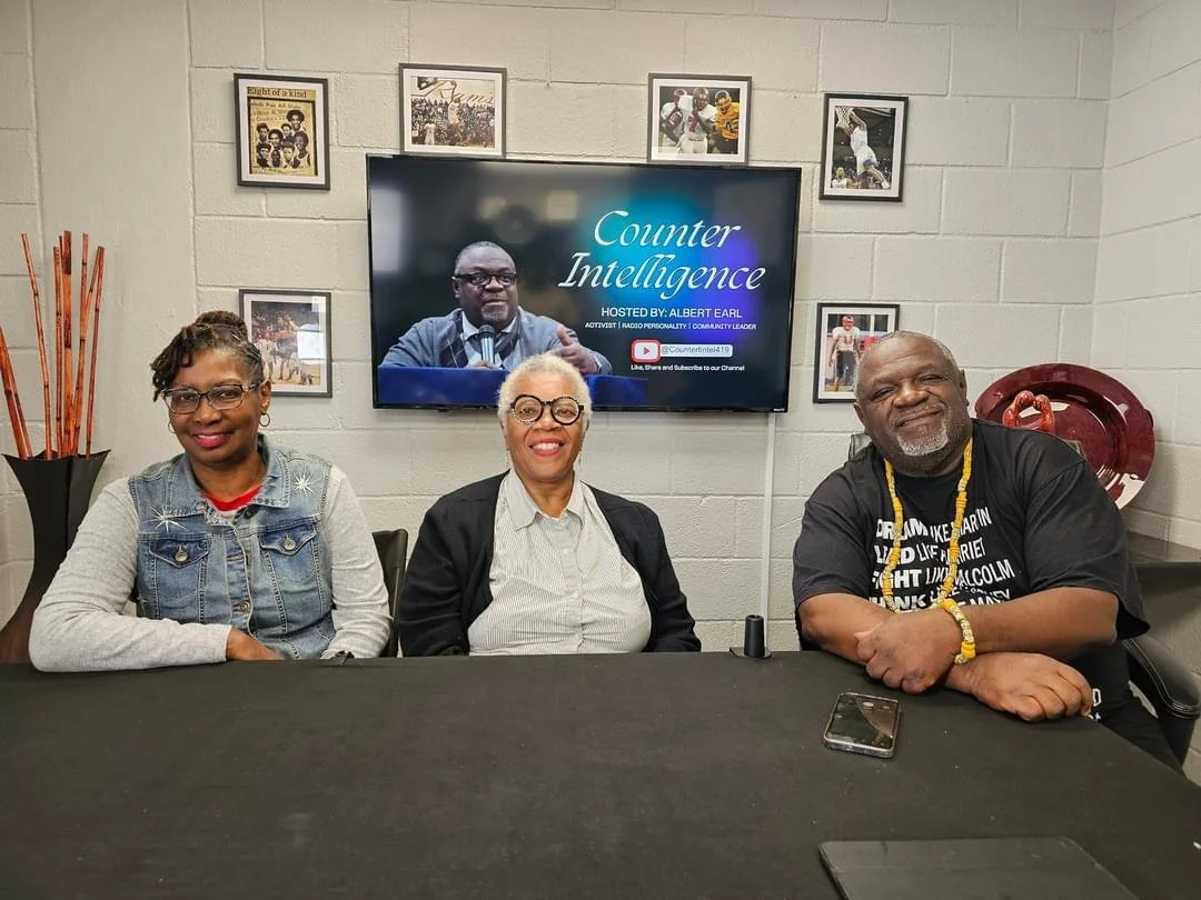 Three people sitting at a table with a TV behind them displaying the words 'Counter Intelligence' and a picture of a man speaking into a microphone. The man is hosted by Albert Earl. The room has framed photos on the wall and a decorative vase with sticks on the left.