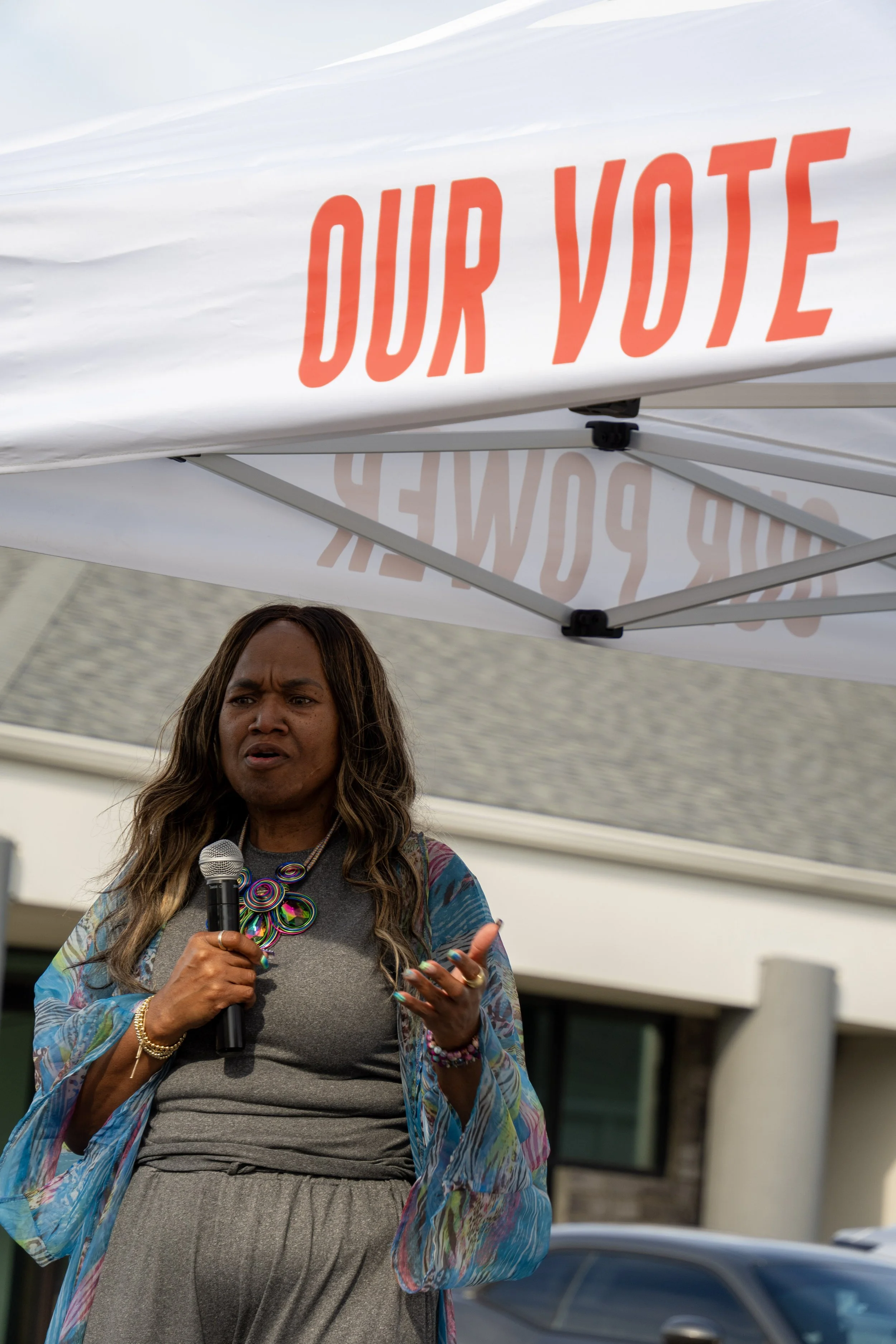 Woman speaking into a microphone under a white canopy with red text that says "OUR VOTE".