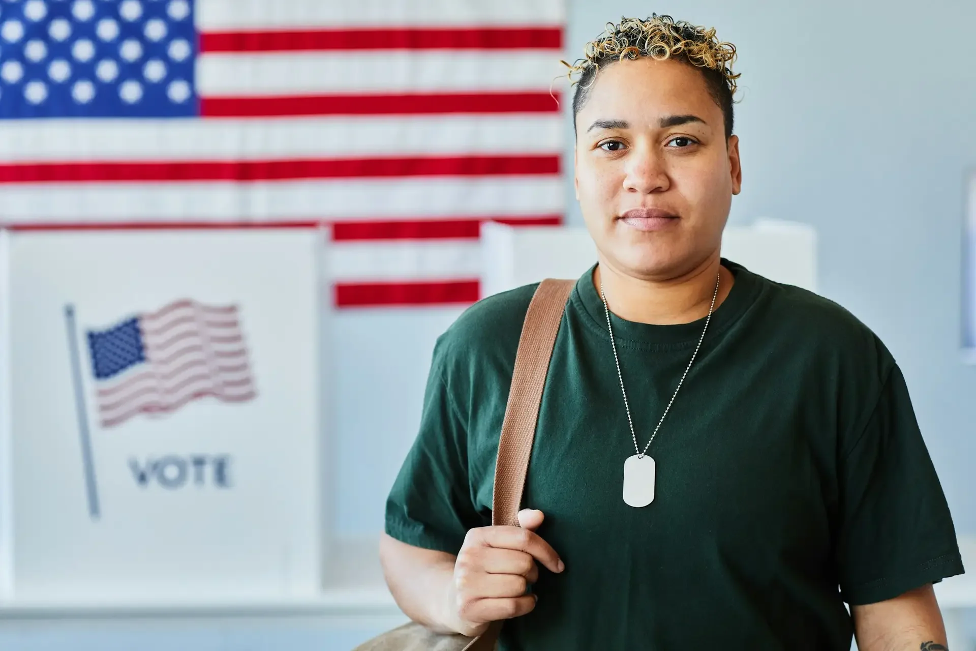 Young man with short, curly, blonde-tipped hair, wearing a green T-shirt and dog tag necklace, standing in front of American flags and voting booths.
