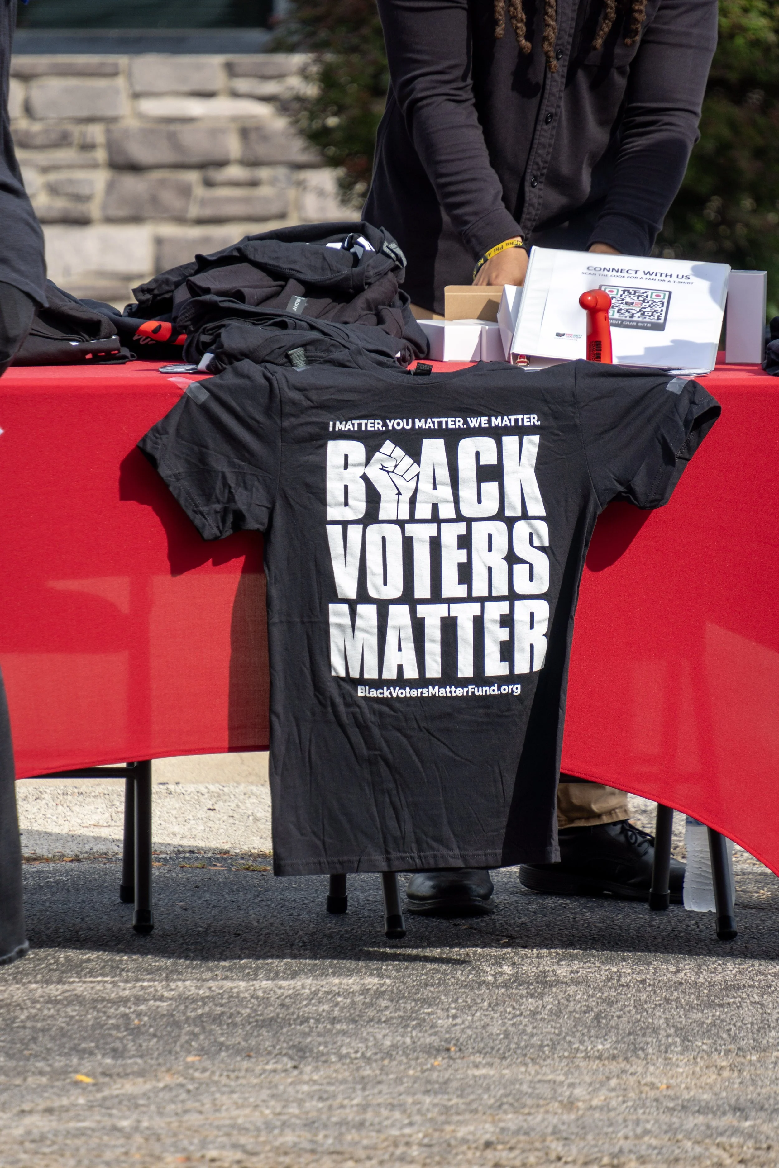 Table with black t-shirt reading 'Black Voters Matter,' backpacks, and a QR code sign at an outdoor event.