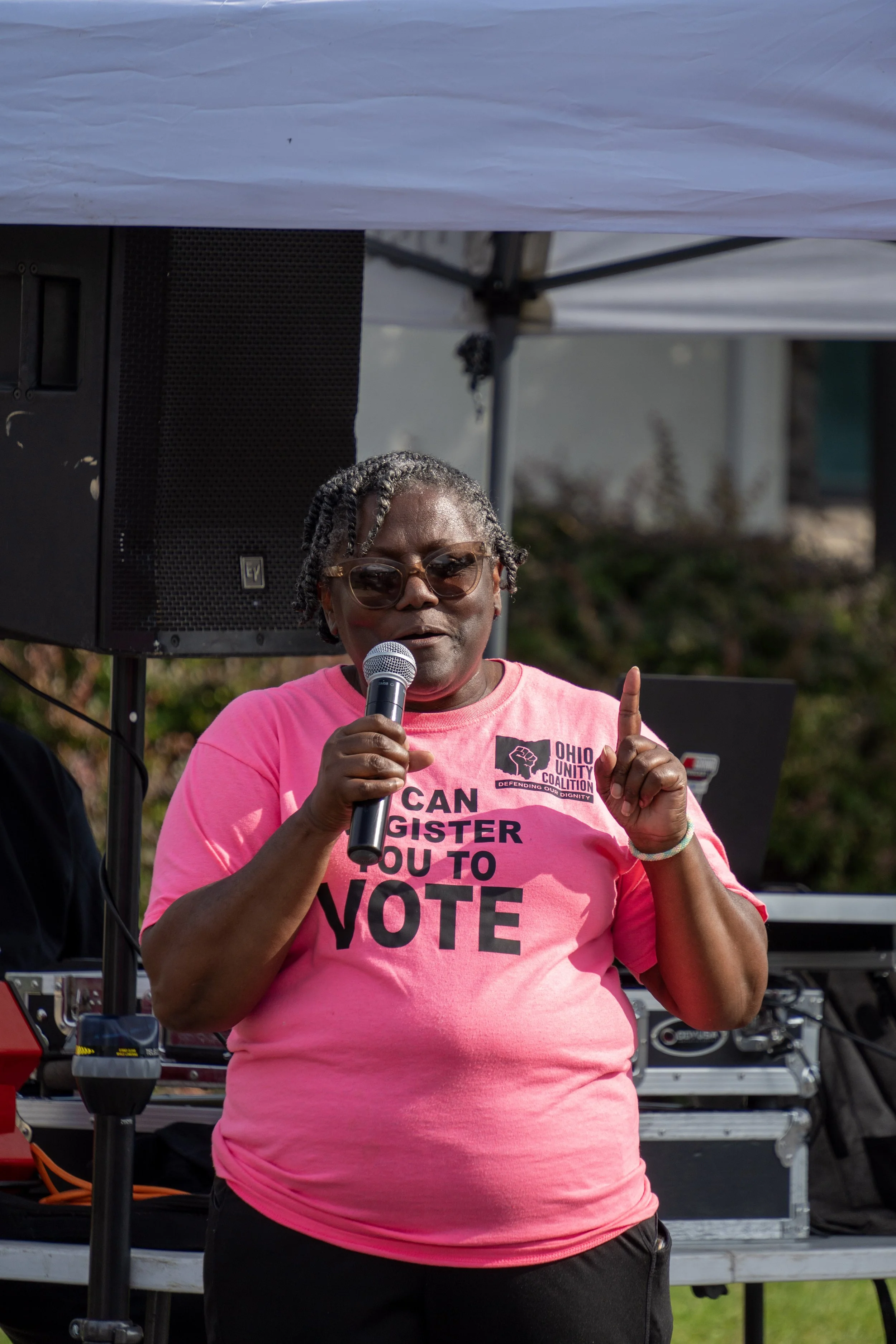 A woman speaking into a microphone at an outdoor event, wearing a pink T-shirt with the message 'CAN REGISTER YOU TO VOTE' and a logo for Ohio Unity Coalition, with large speakers and a stage setup in the background.