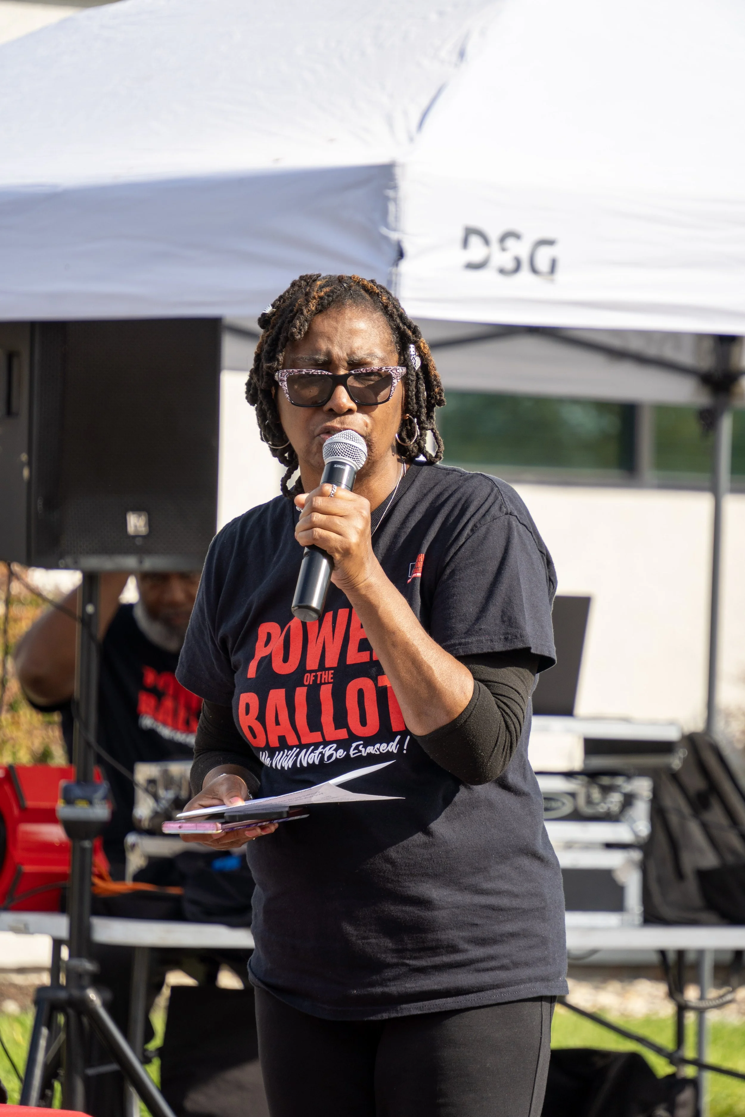 A woman with dreadlocks and glasses speaking into a microphone at an outdoor event, wearing a black t-shirt with red text referencing the power of the ballot. In the background, there is a person at a keyboard and other sound equipment, covered by a 
