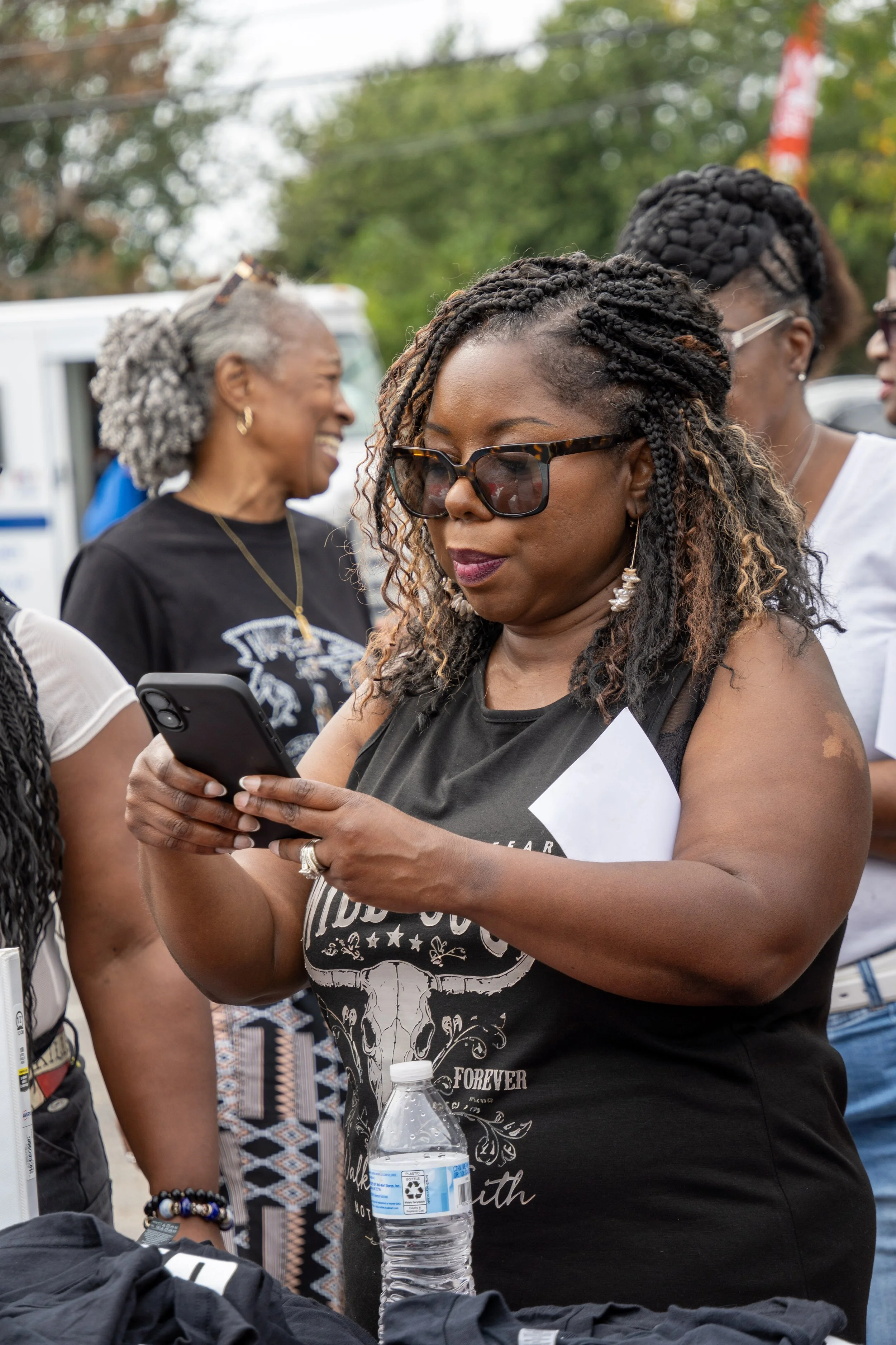 A woman with braided hair and sunglasses looking at her phone at an outdoor gathering. Several people are in the background, smiling and interacting.