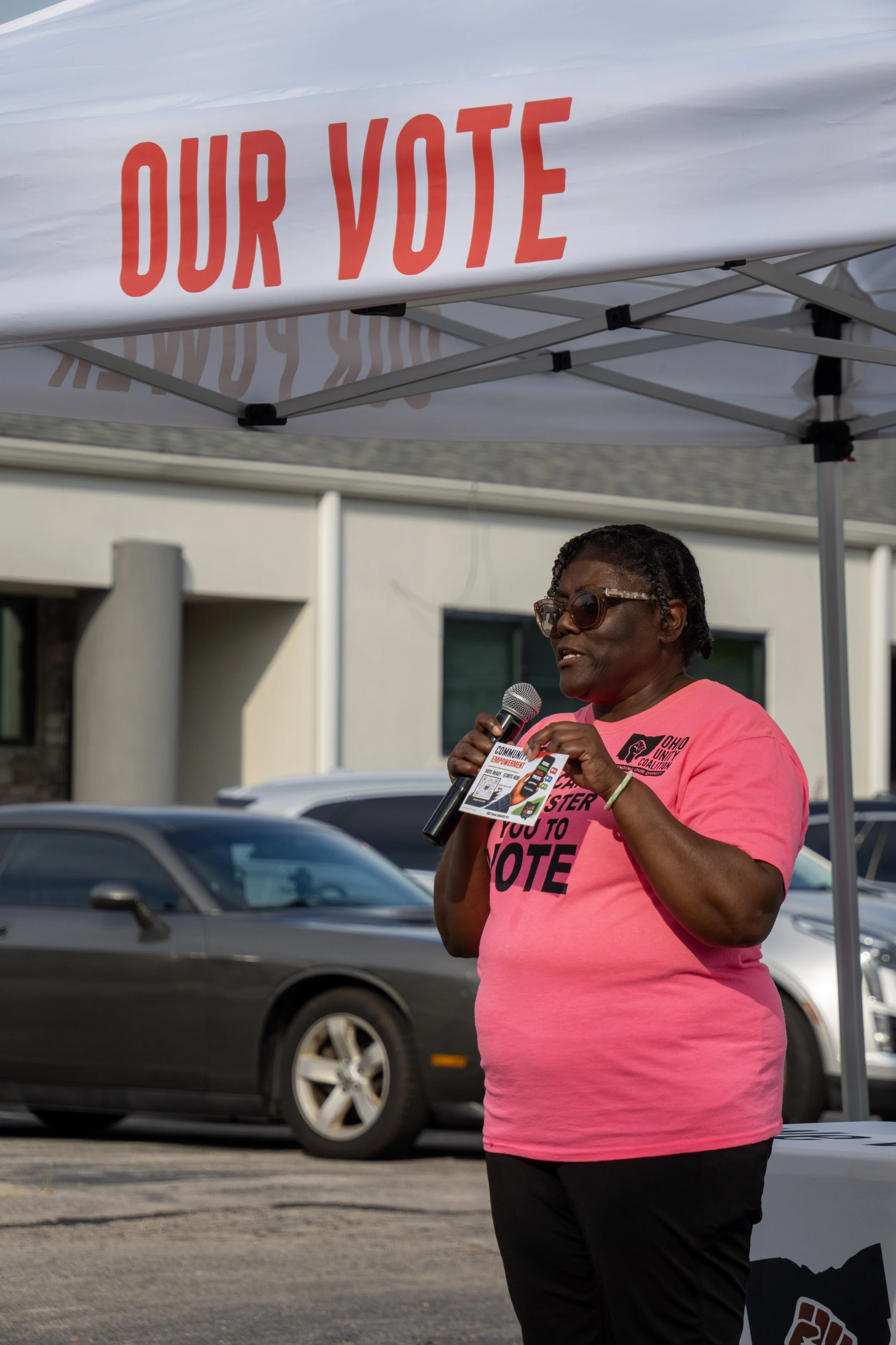 A woman wearing sunglasses and a pink T-shirt, holding a microphone and standing under a white canopy with red lettering that reads 'OUR VOTE.' Cars are visible in the background, indicating an outdoor setting.