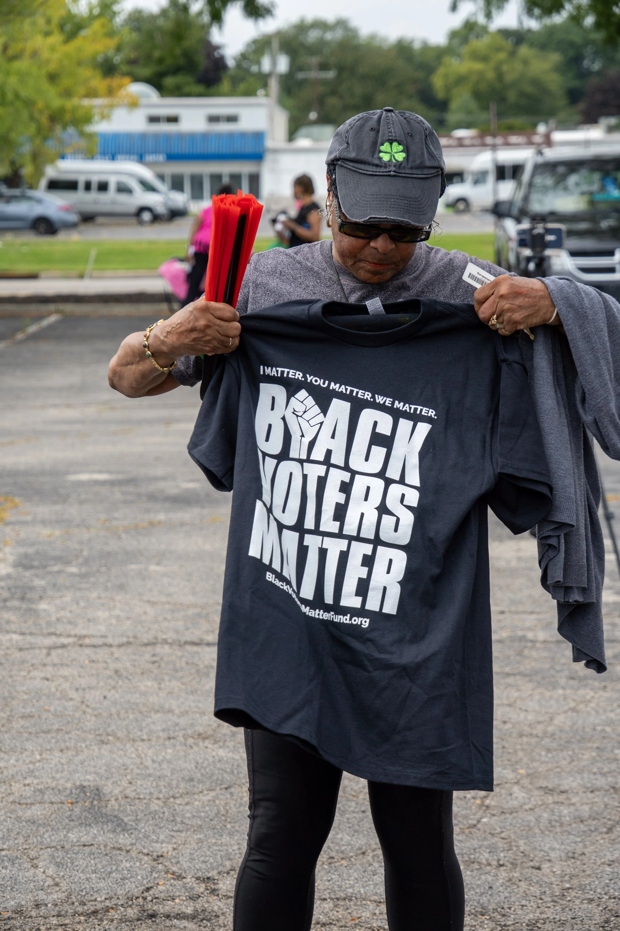 A woman in black leggings and a dark T-shirt holding a black 'Back the Voters' T-shirt and a bundle of red flyers, standing outdoors in a parking lot with people and cars in the background.