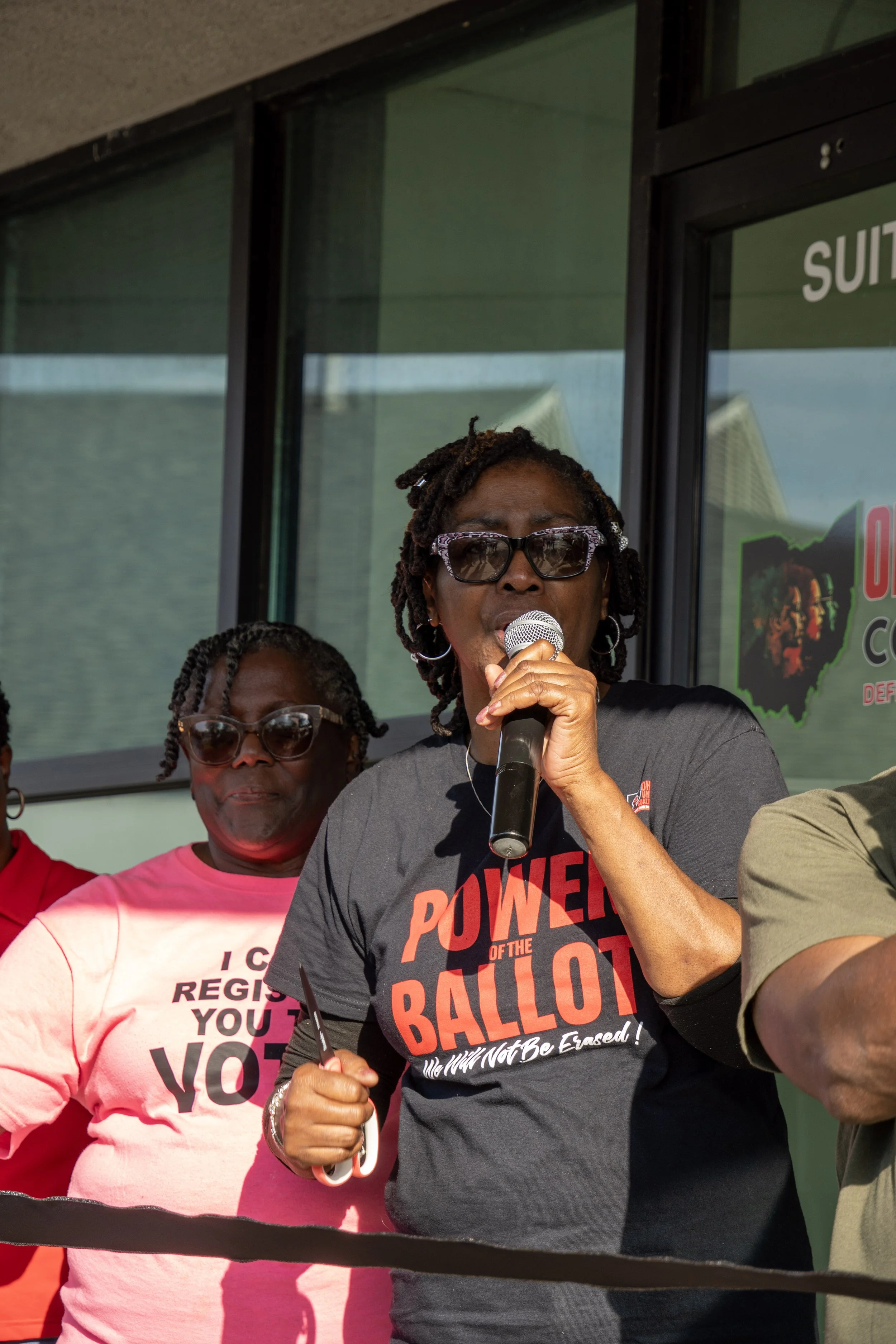 A group of women participating in a rally or protest, with one woman speaking into a microphone. The woman in front is wearing sunglasses and a black T-shirt with red text about voting and democracy. A woman in background wears a pink T-shirt support
