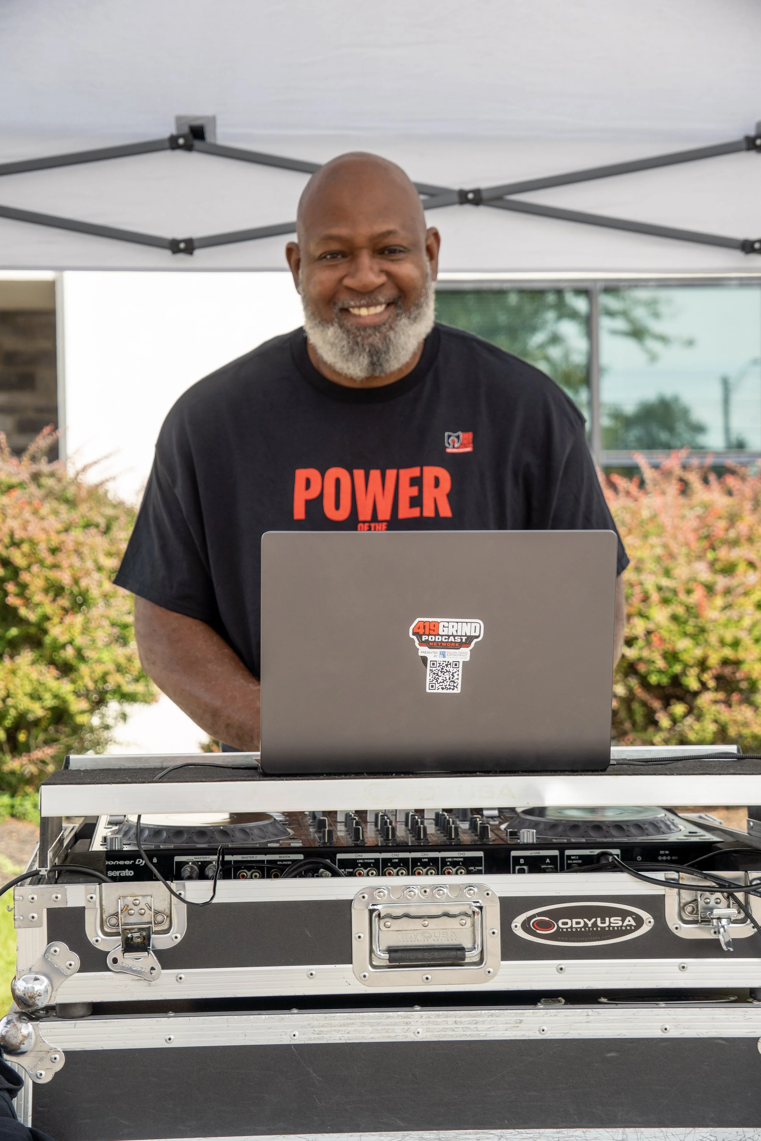 A middle-aged man with a gray beard smiling at a DJ setup outdoors, wearing a black T-shirt with red and white text, and working at a laptop with a DJ mixer in front of him.