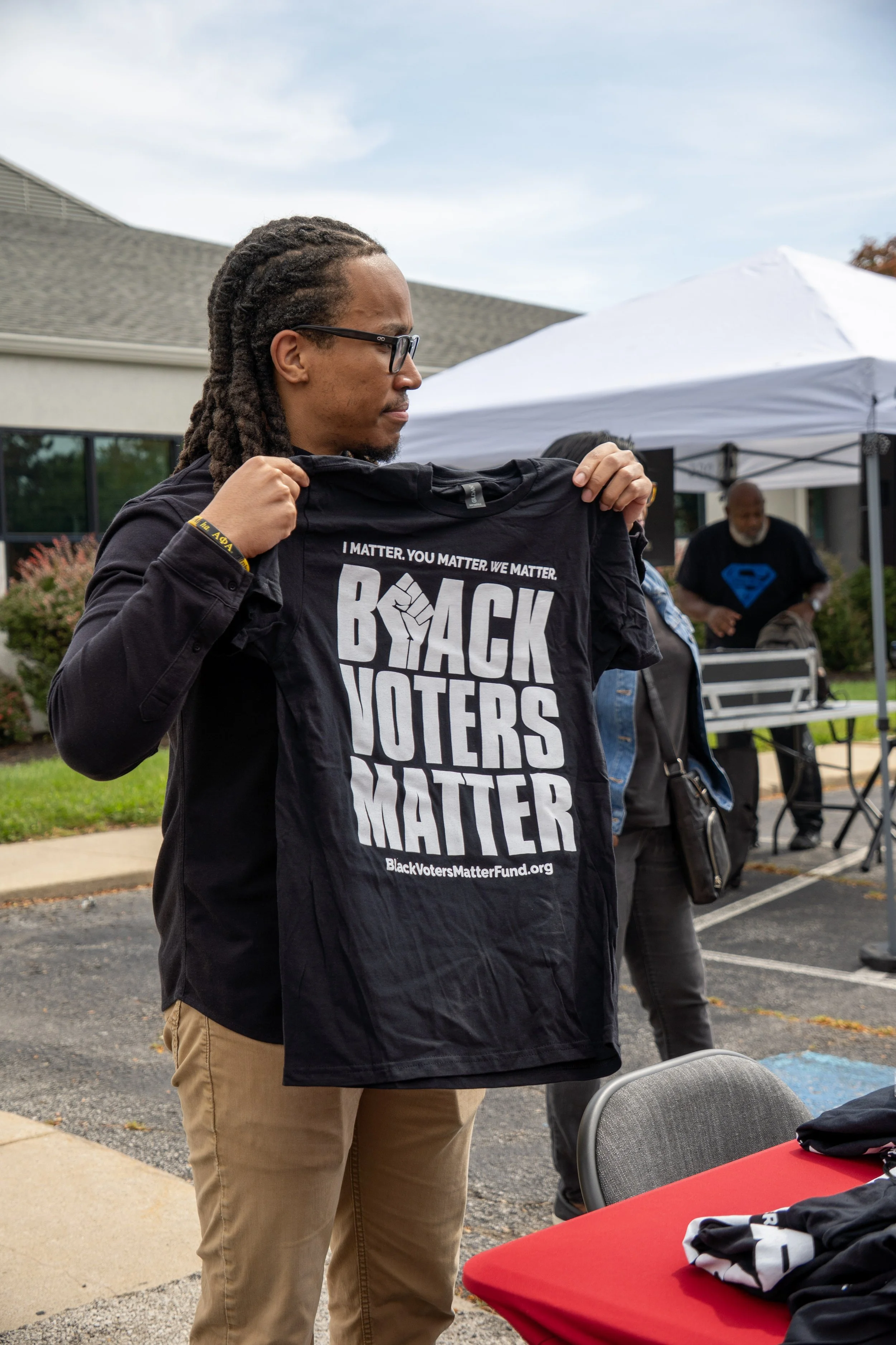 A man with long dreadlocks, wearing glasses, holding a black T-shirt that reads 'BACK THE VOTERS MATTER' at an outdoor event. There is a white canopy tent and other people in the background.