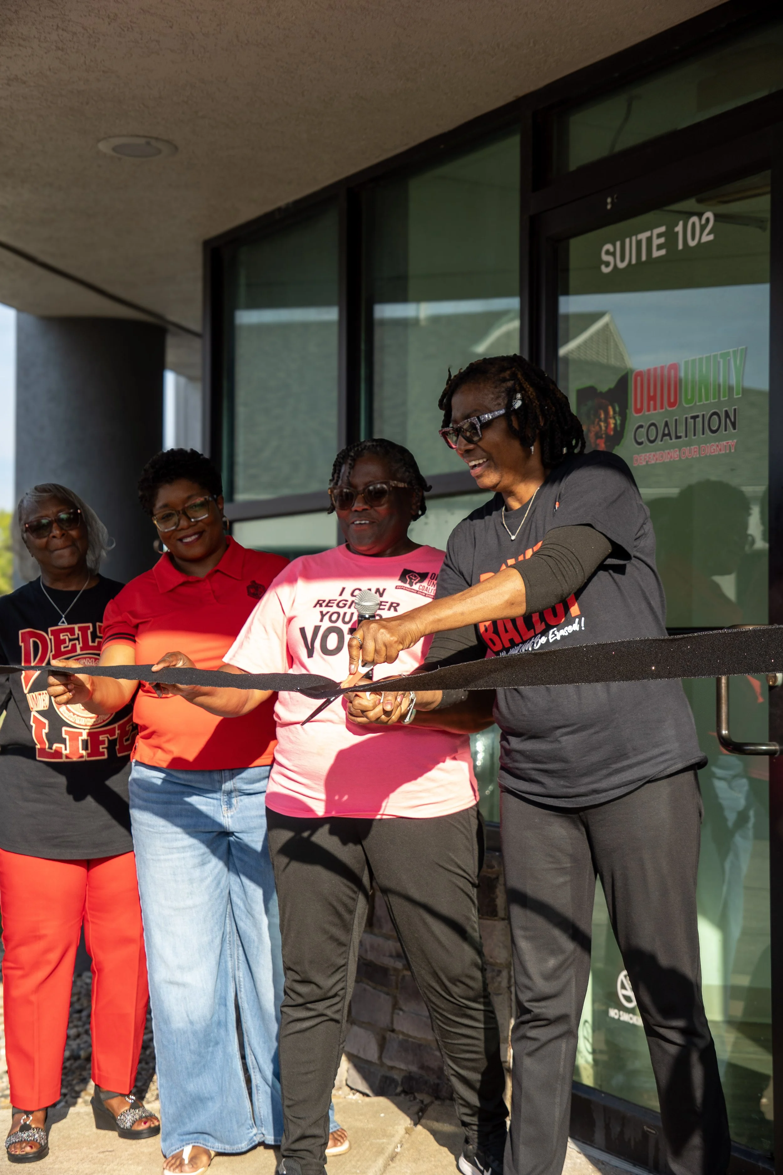 Four women standing together outside a building with a sign for Ohio Unity Coalition; one woman is cutting a ribbon for an event.