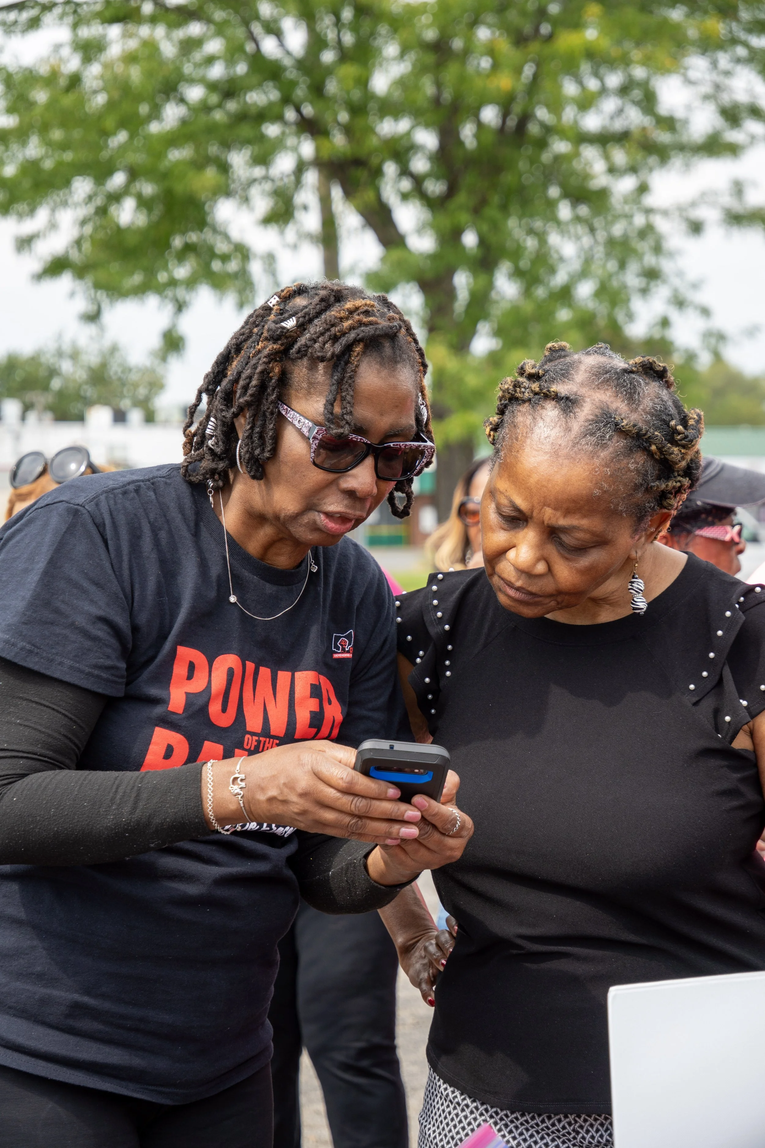 Two women looking at a phone together outdoors, one wearing glasses and a black shirt with red text, and the other wearing earrings and a black top, with green trees in the background.
