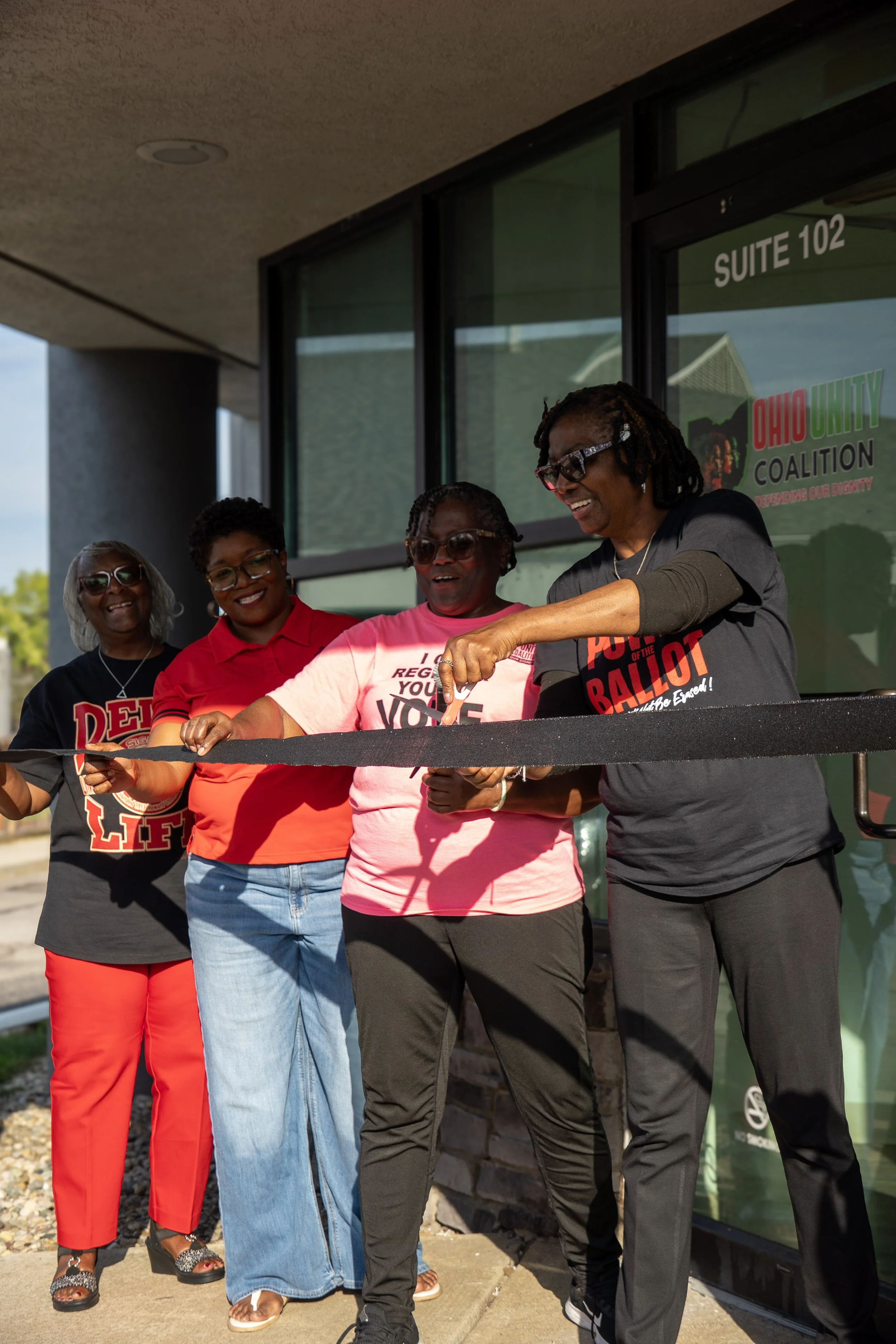A group of four women standing together outside a building with a sign that reads 'Suite 102' and 'Ohio Unity Coalition' in the background. They are smiling, with one woman cutting a ribbon in front of a glass door.