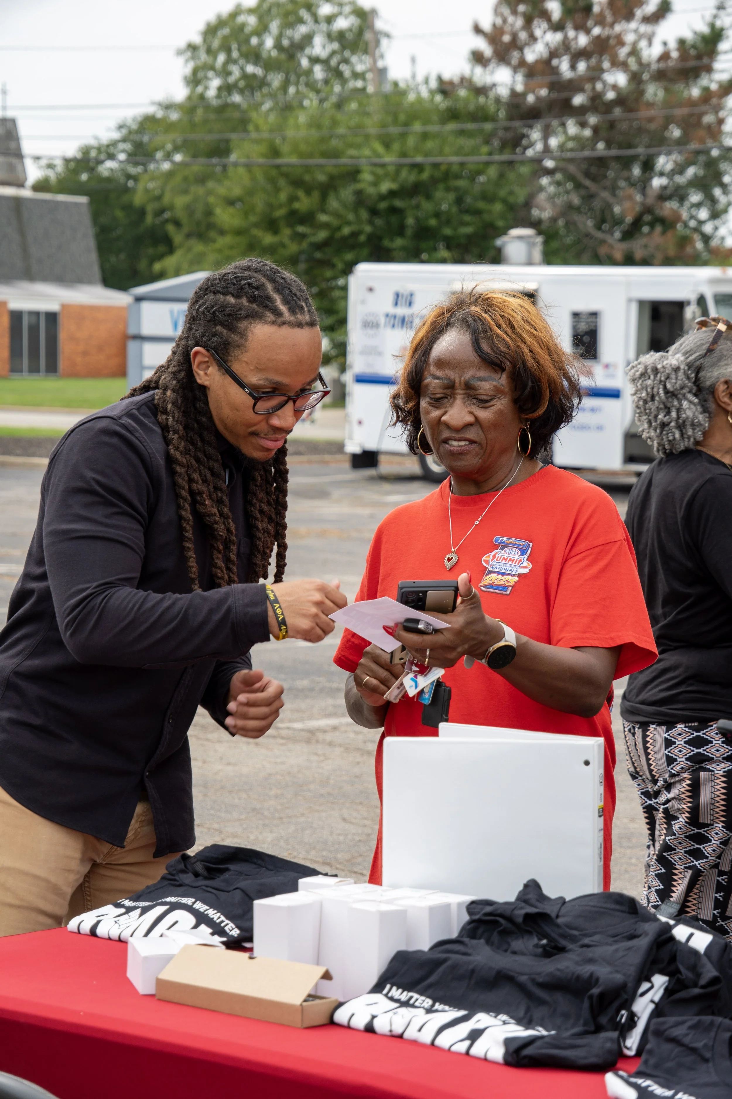A man with long dreadlocks and glasses and a woman with short hair and hoop earrings are looking at a phone together. They are standing at a table with some items on it, including T-shirts with the slogan 'Black Lives Matter' and a white box. In the background, there is a mobile unit and some trees.