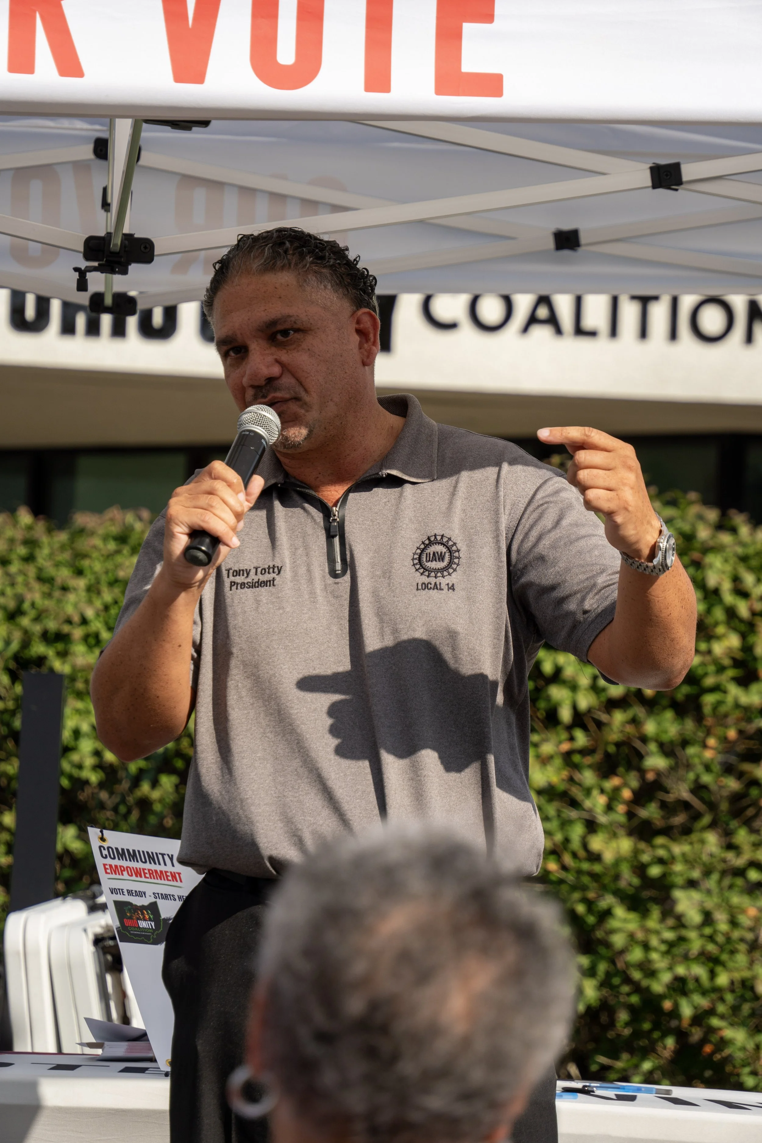 Man speaking into a microphone at a rally or gathering under a white tent with red and black signage, wearing a gray polo shirt with a UAW logo and the name Tony Totty.