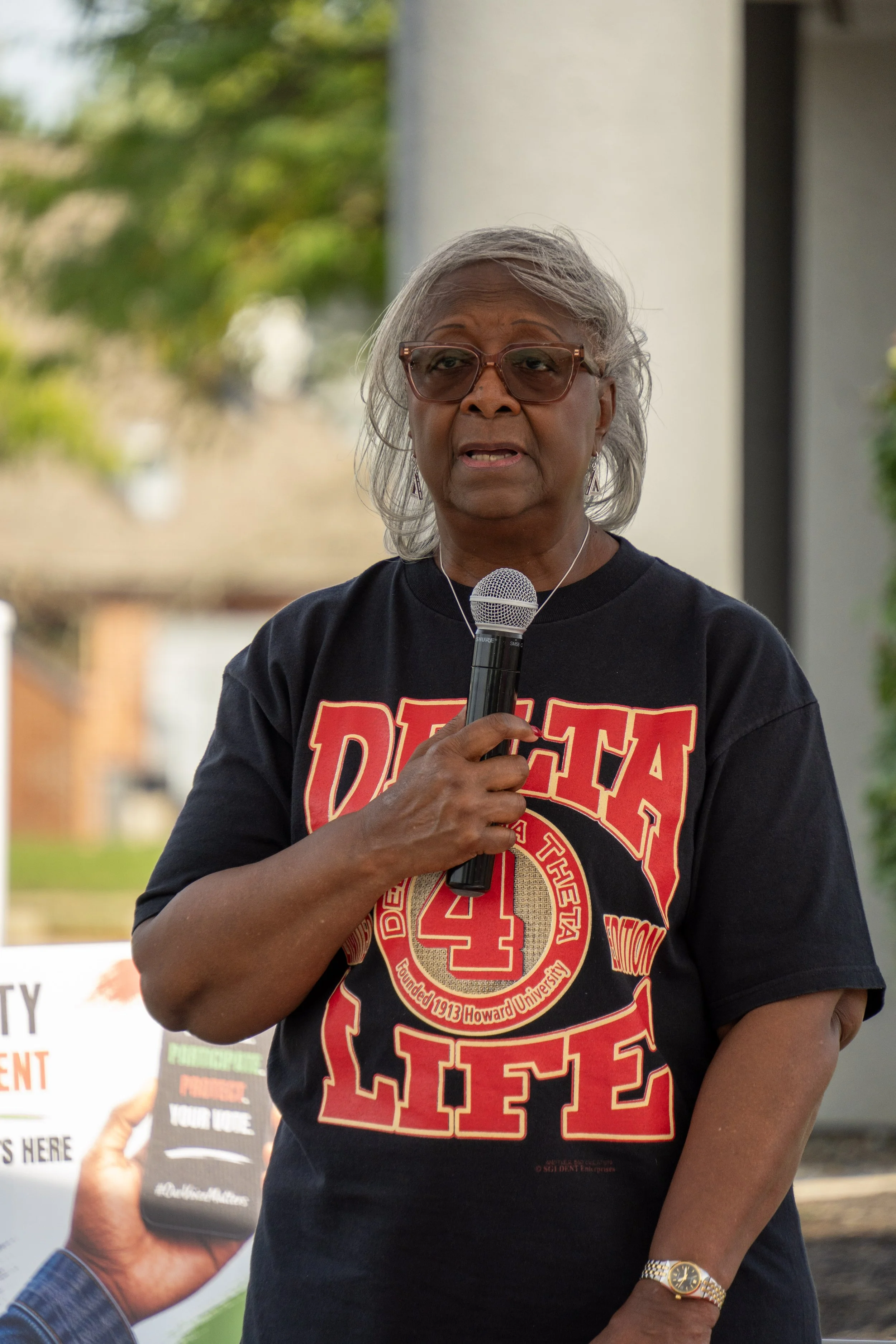 An elderly woman with gray hair and glasses speaking into a microphone during an outdoor event, wearing a black T-shirt with red and gold lettering that reads 'DETA,' '4,' and 'Howard University,' and a gold wristwatch.