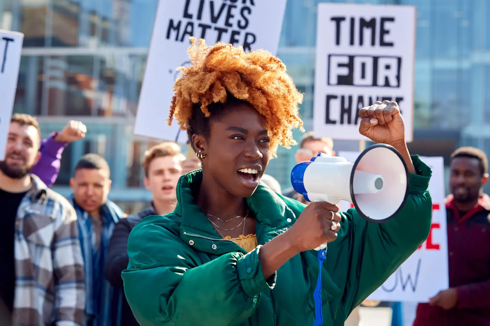A woman with curly orange hair holding a megaphone and raising her fist at a protest, with signs in the background that read 'LIVES MATTER' and 'TIME FOR CHANGE', amid a crowd of protesters.