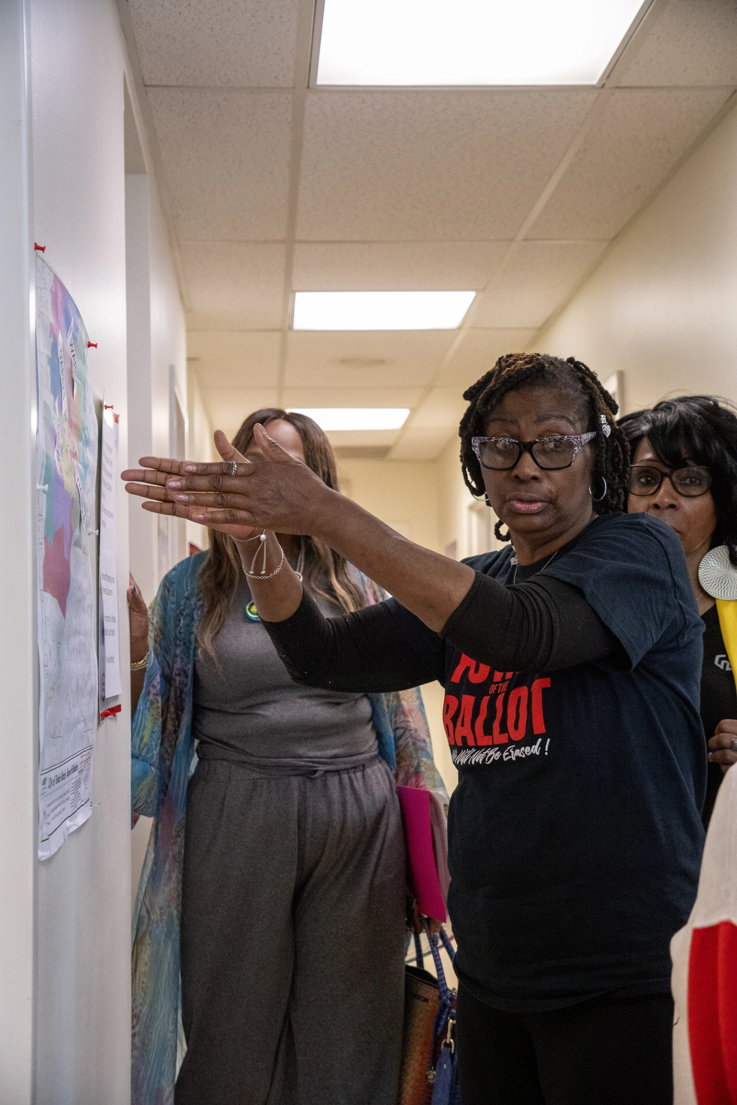 A group of women in an indoor setting, with one woman in the foreground making a gesture with her hand while wearing glasses and a black T-shirt with red and white lettering. Others are partially visible in the background.