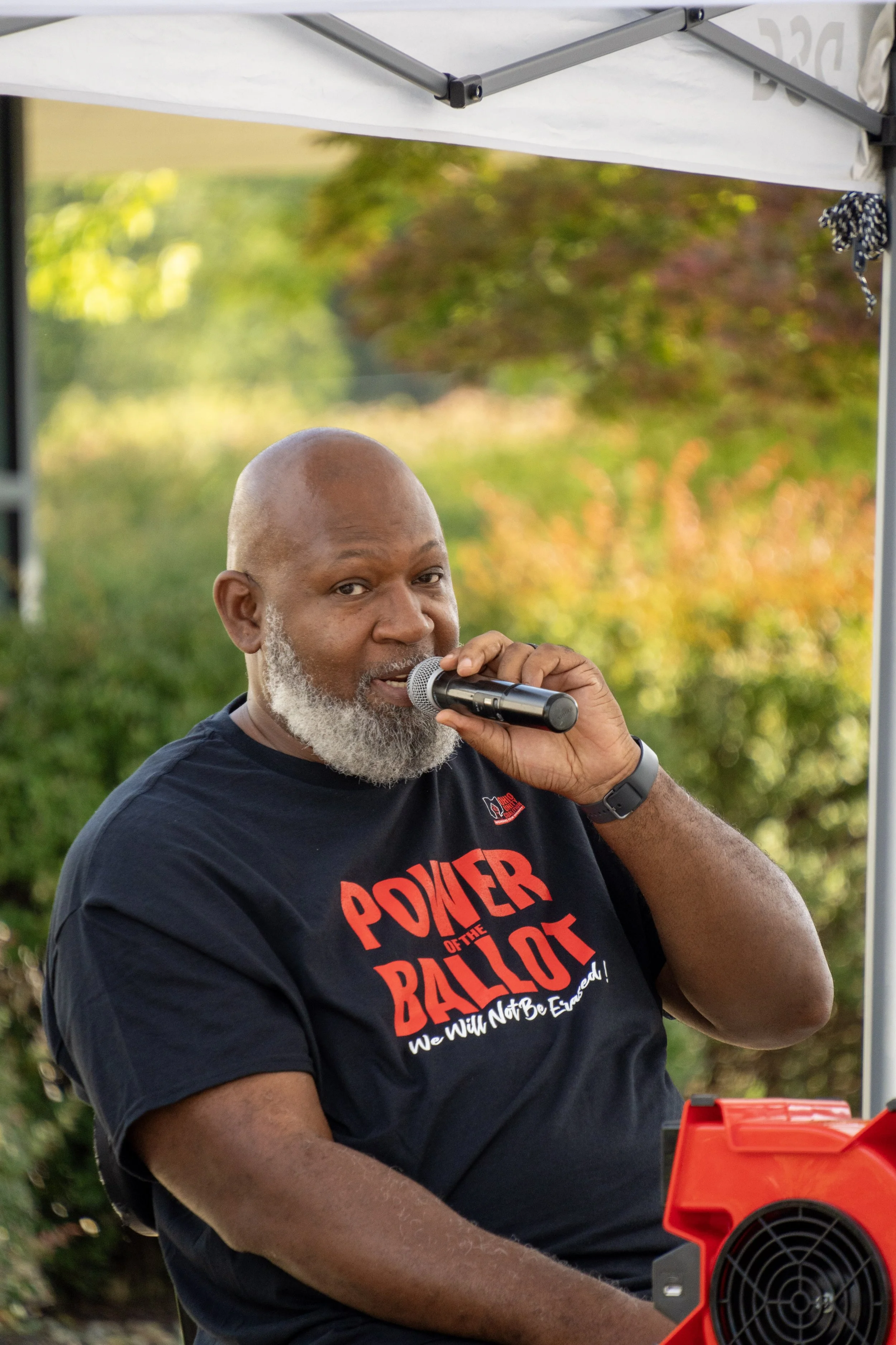 A man with a gray beard speaking into a microphone outdoors, wearing a black t-shirt with red and white text, standing under a white tent with orange and red equipment in the foreground, with trees and bushes in the background.