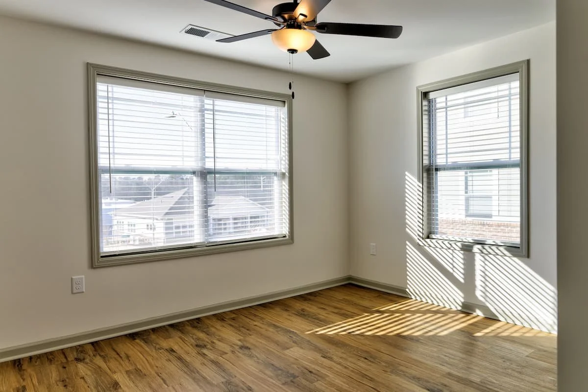 Empty room with two windows with blinds, a ceiling fan with a light, and wood flooring, sunlight casting shadows through the blinds at Birch & Branch Apartments in Raleigh, NC.