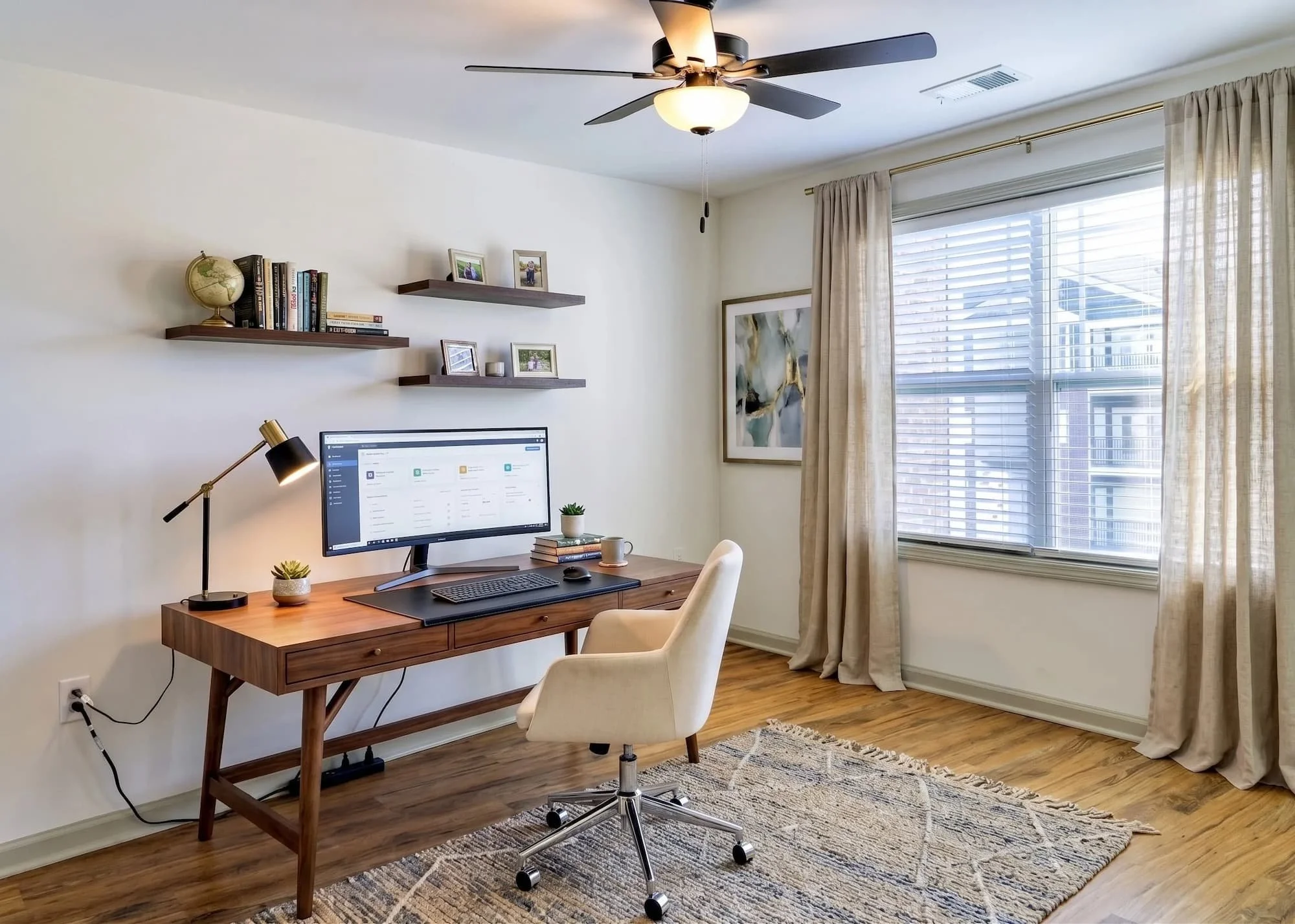A home office at Birch and Branch apts with a wooden desk, a beige swivel chair, a computer monitor, small potted plants, and framed photographs on wall-mounted shelves. There's a large window with blinds and beige curtains, a ceiling fan with lights