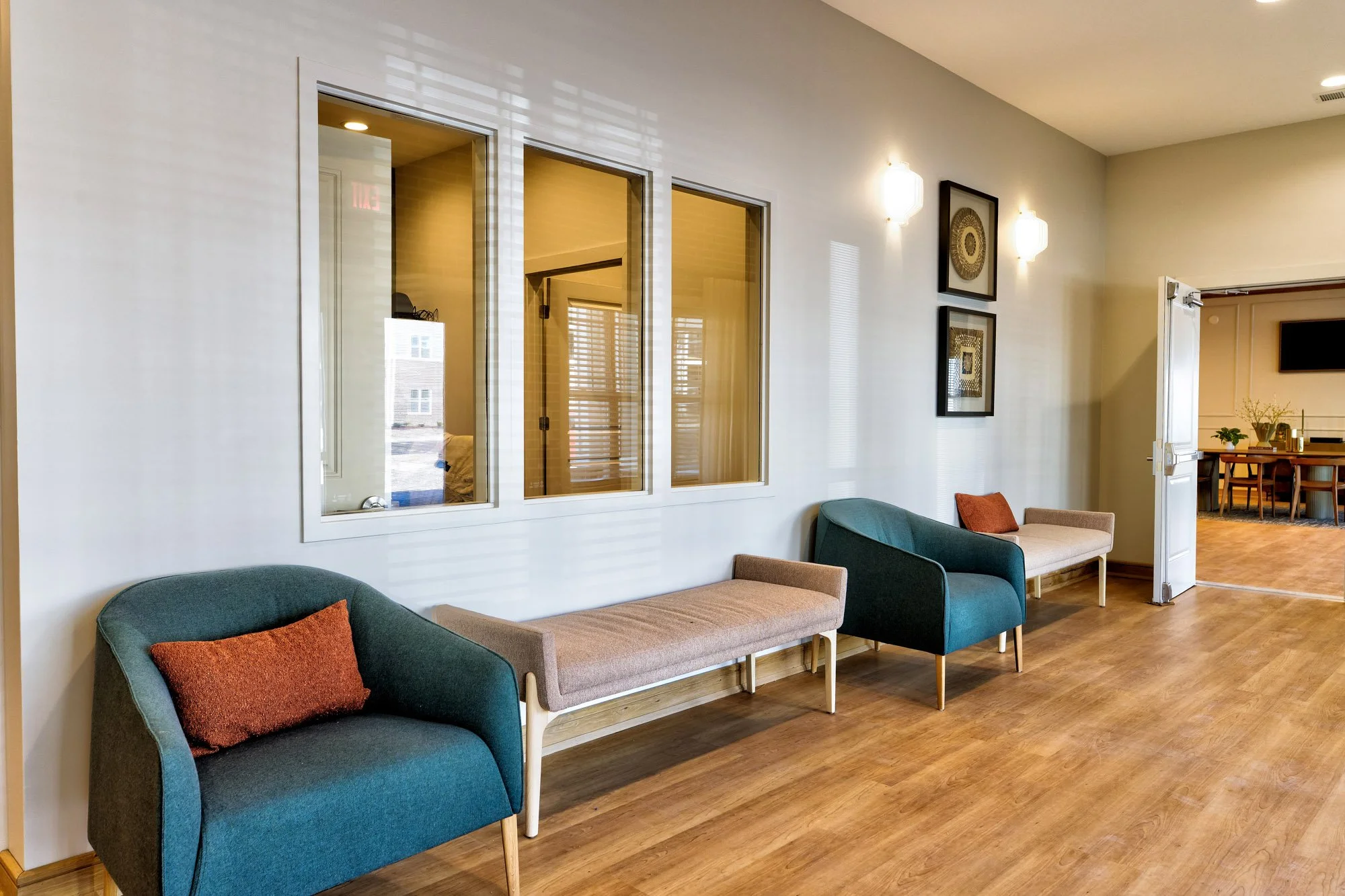 Communal waiting area at Birch & Branch Apartments with a teal armchair with a red pillow, a white wall with framed circular art, and outdoor view through windows. An open door reveals a dining area with a wooden table, chairs, and indoor plants.